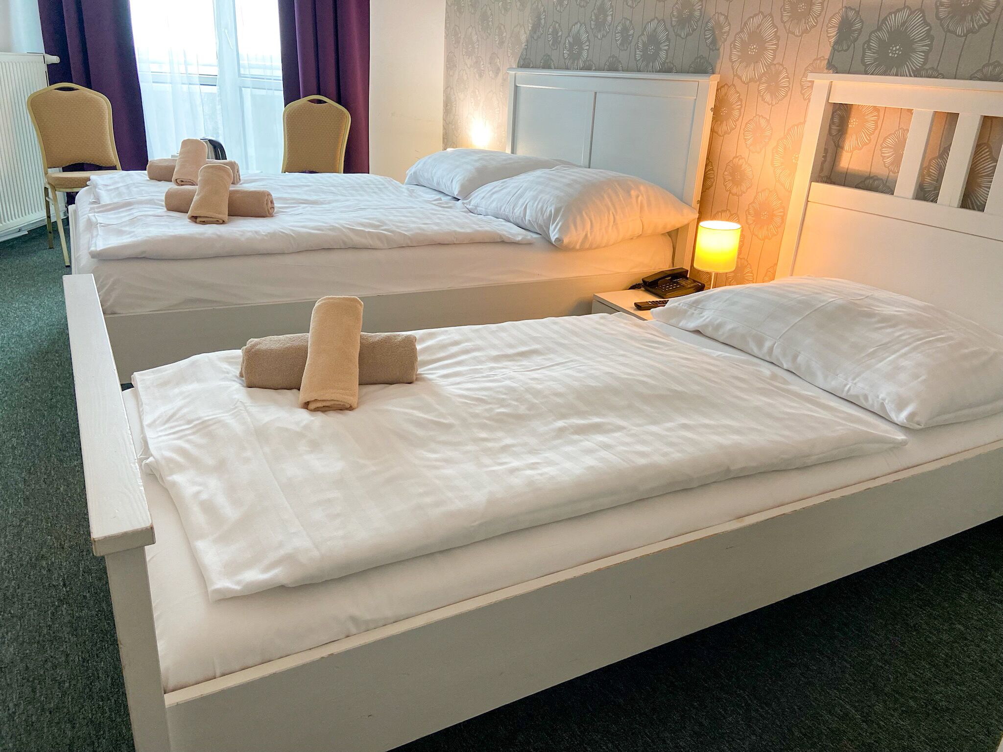 Een hotelkamer met twee eenpersoonsbedden, elk voorzien van witte lakens en handdoeken, een nachtkastje met een lamp en een hoofdbord met een bloemenprint.