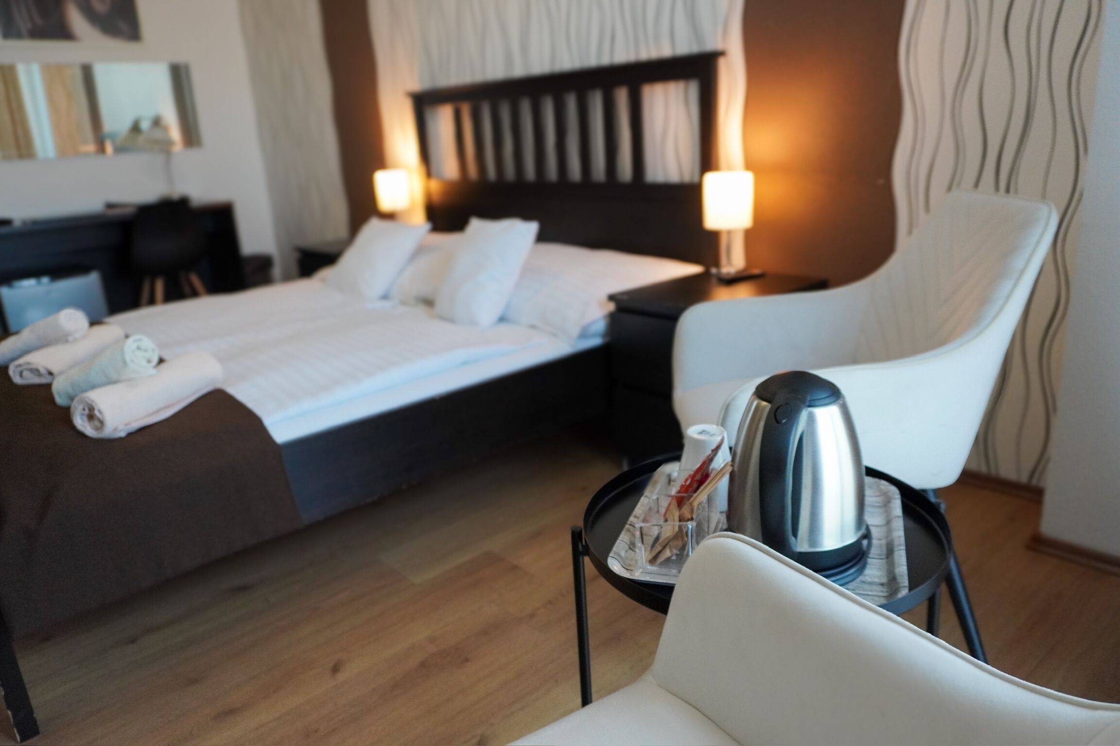 A hotel room with a neatly made bed, a sitting area with a chair and a small table, and a bedside table with a kettle.