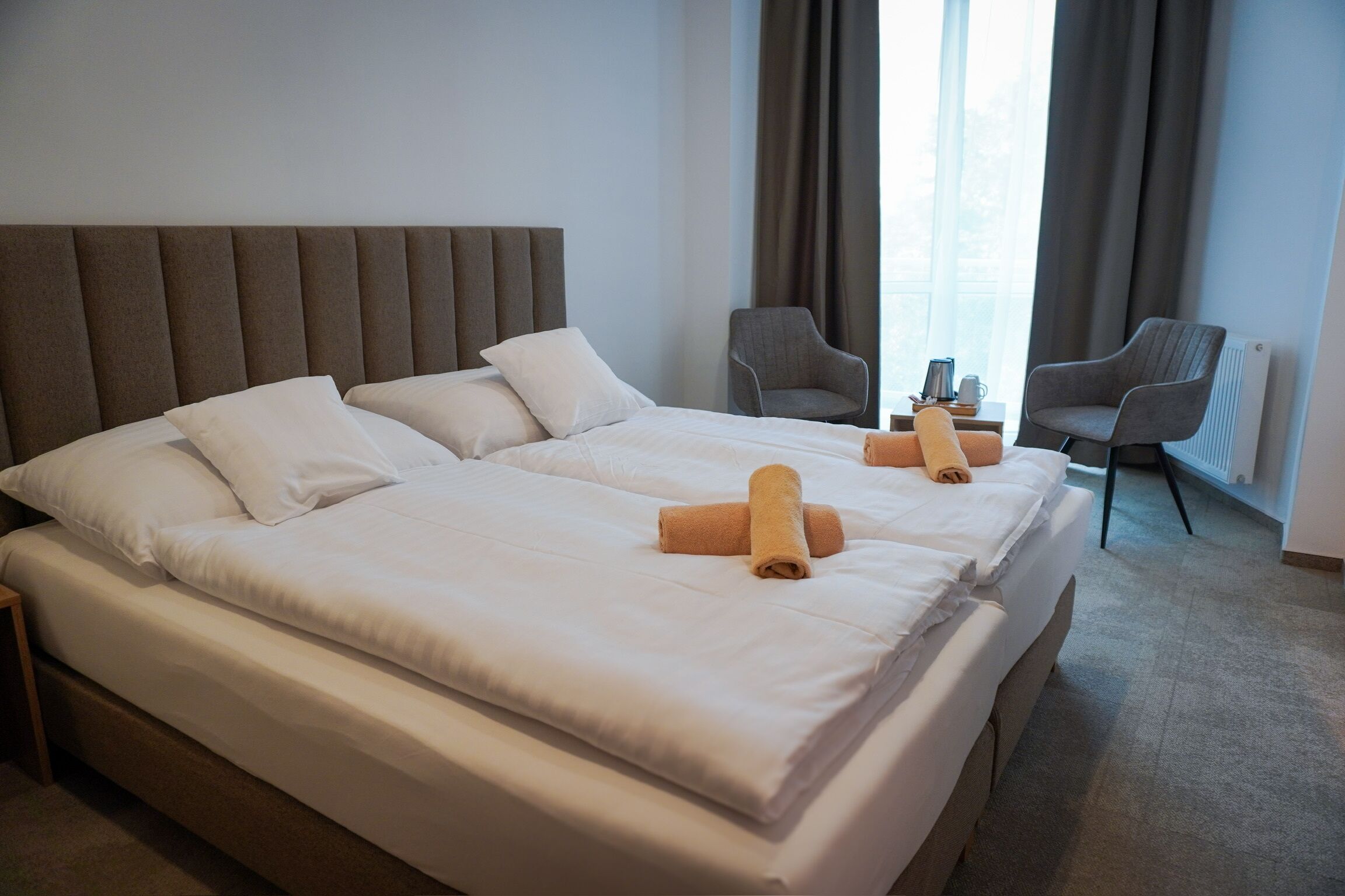 A double bed with white linens and two rolled towels, a grey upholstered headboard, two grey armchairs, and a window with curtains.