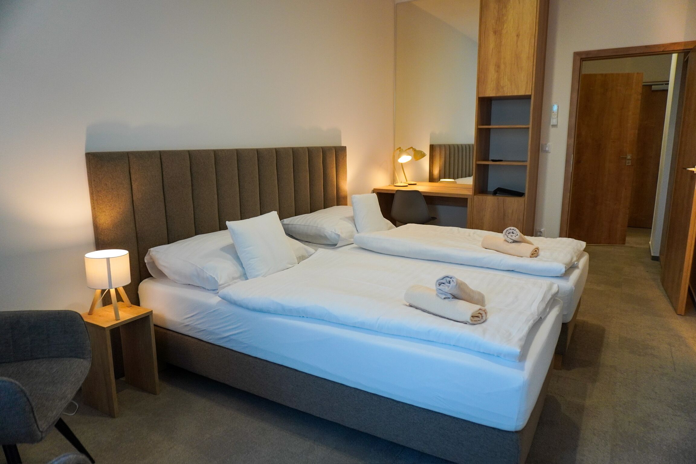 A hotel room with a large bed, a bedside table with a lamp, a chair, and a wooden wardrobe.