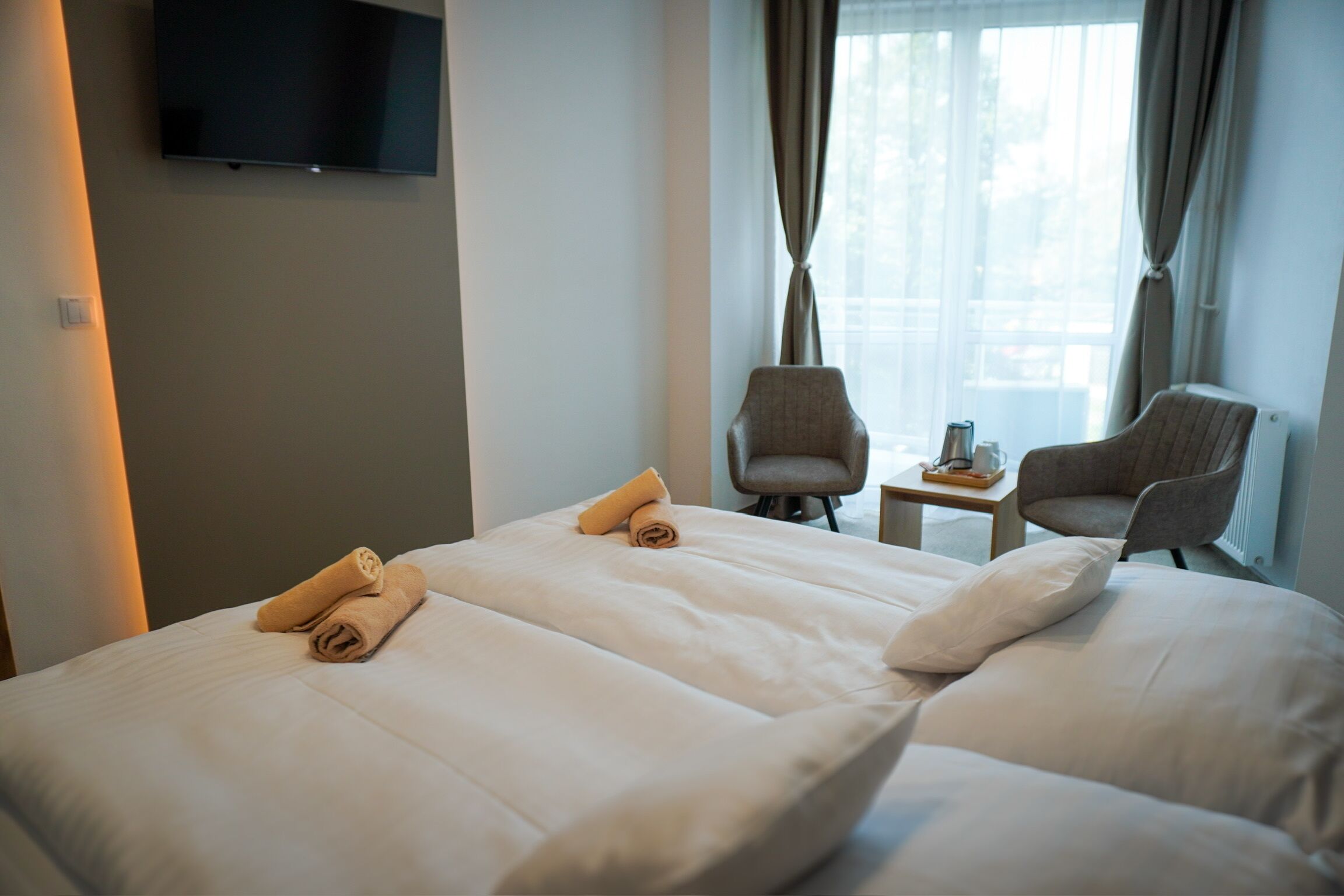 Habitació d'hotel amb dos llits, un televisor penjat a la paret i dues cadires a prop d'una finestra amb cortines.