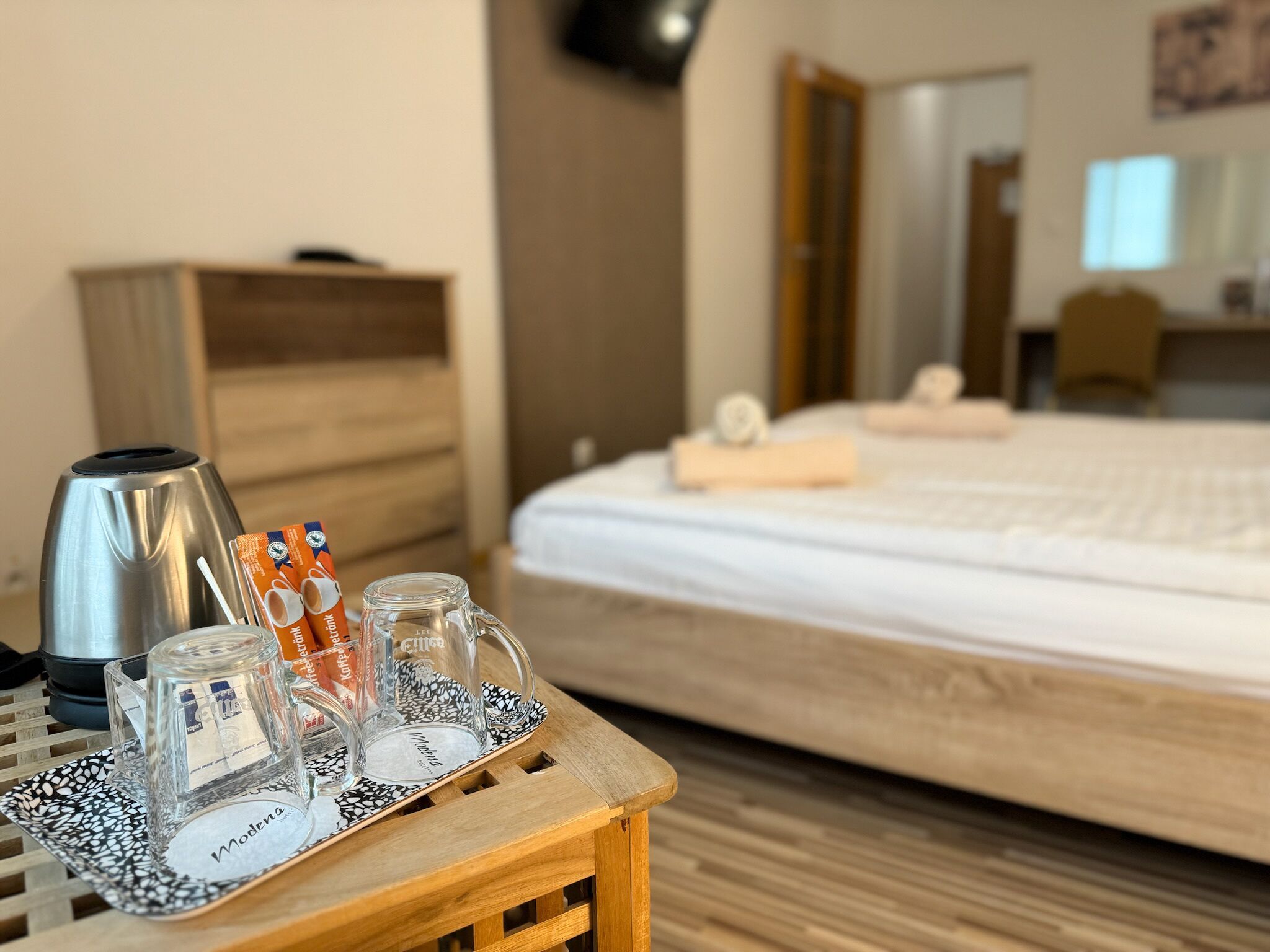 A hotel room with a bed, a wooden nightstand with a coffee maker, a tray with cups and a glass, and a small table with a tray.