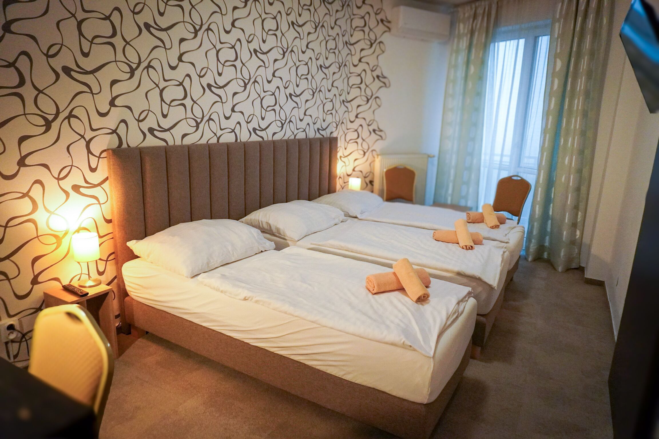 A hotel room with two beds, a headboard, a lamp, a chair, and a window with curtains.