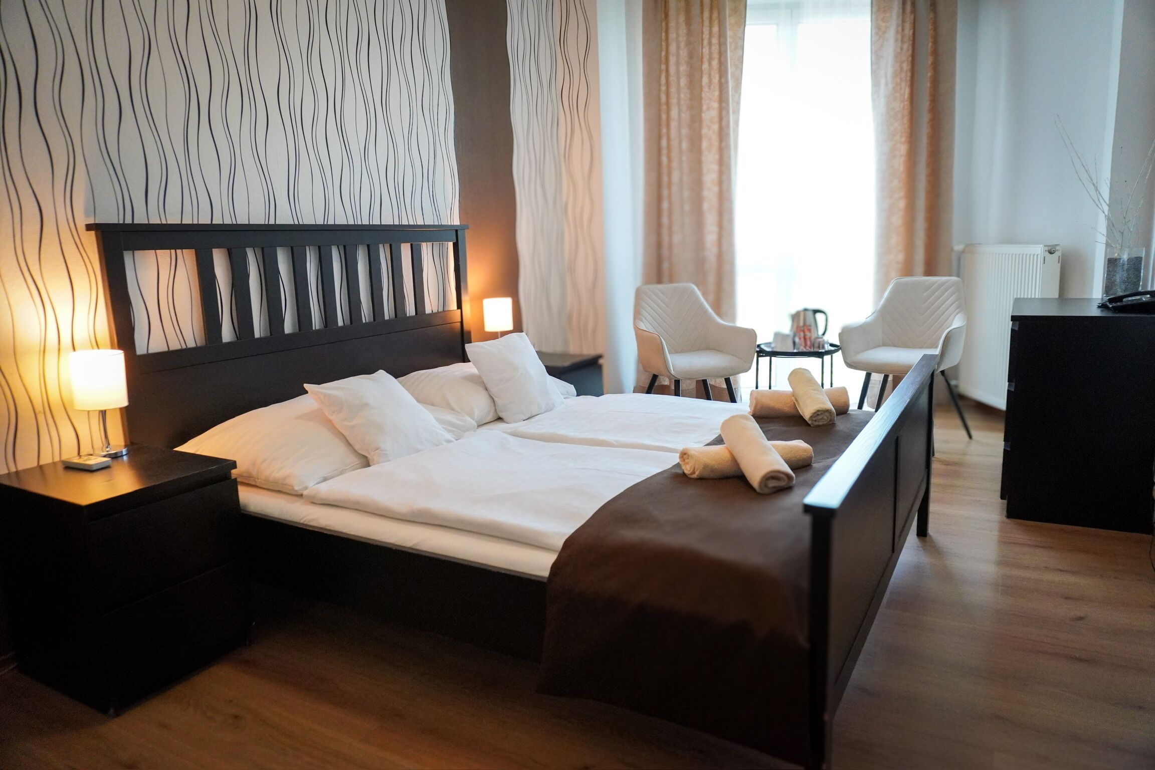 A hotel room with a large bed, two bedside tables, a nightstand, a desk, two chairs, and a radiator.