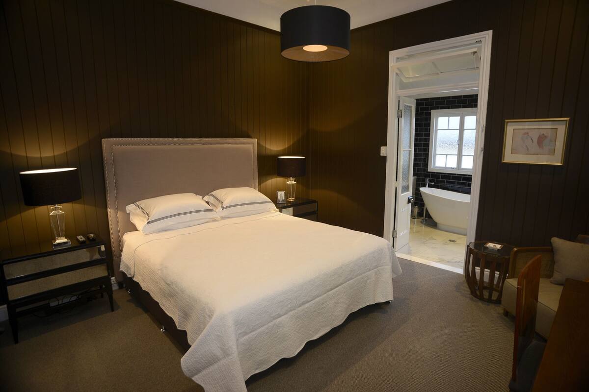 A bedroom with a large bed, bedside lamps, a nightstand, a wooden bench, and a door leading to a bathroom.
