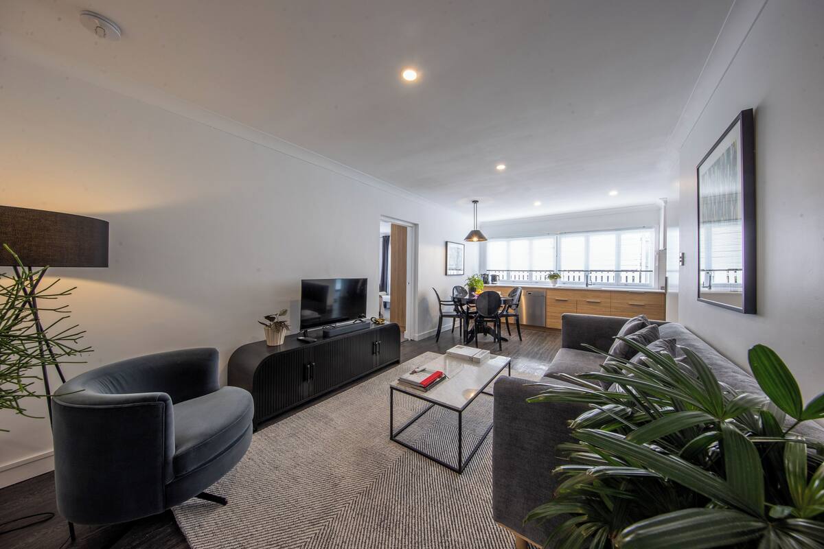 A modern living room with a sofa, armchair, and a coffee table. There is a television on a black cabinet, and a dining area with a table and chairs.