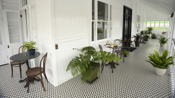 Un largo pasillo con paredes blancas y suelo a cuadrícula en blanco y negro, que conduce a un comedor con una mesa y sillas, y plantas en macetas a lo largo de las paredes.