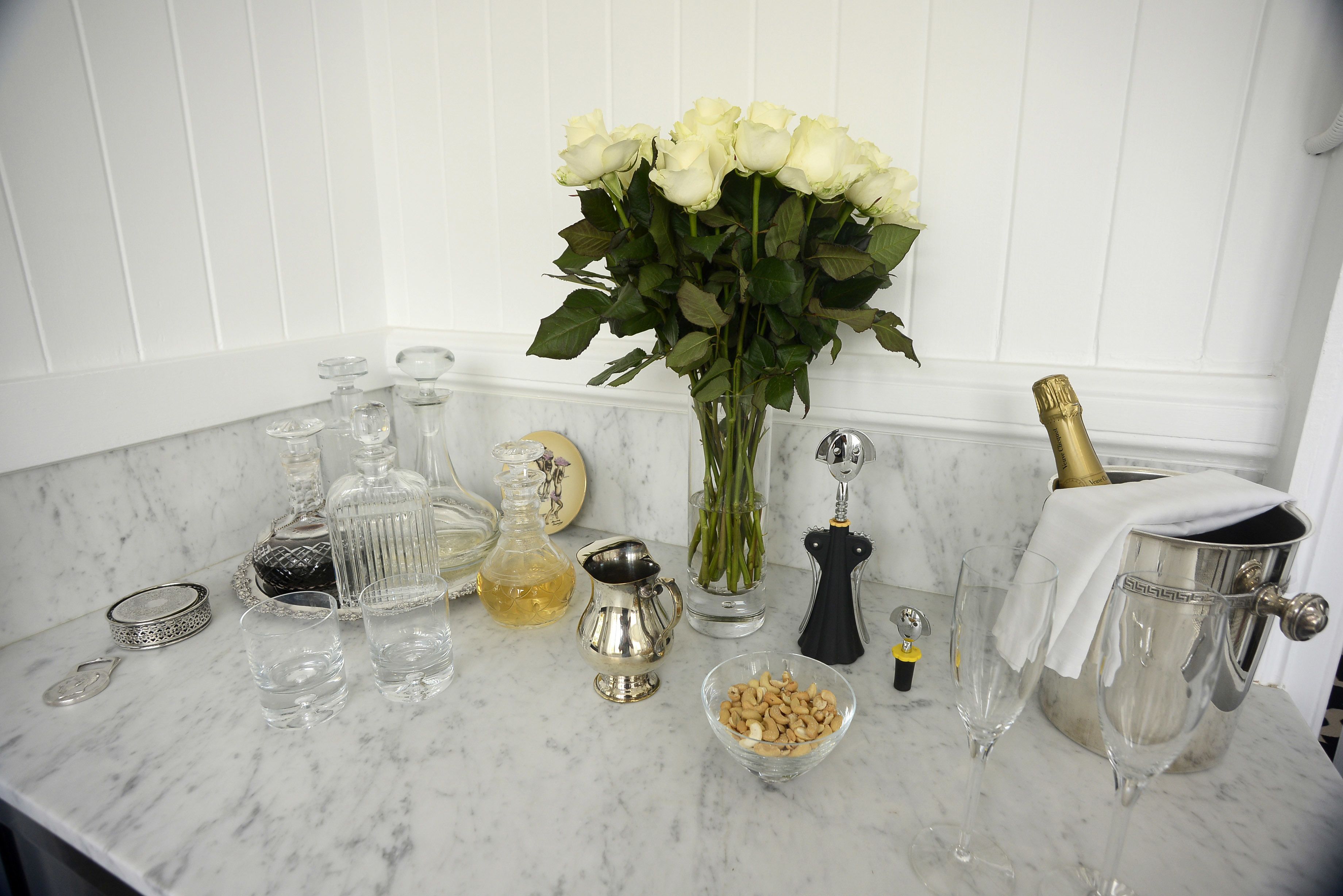 A marble countertop with various glassware, a vase of white roses, a bottle of champagne, and a small bowl of nuts.