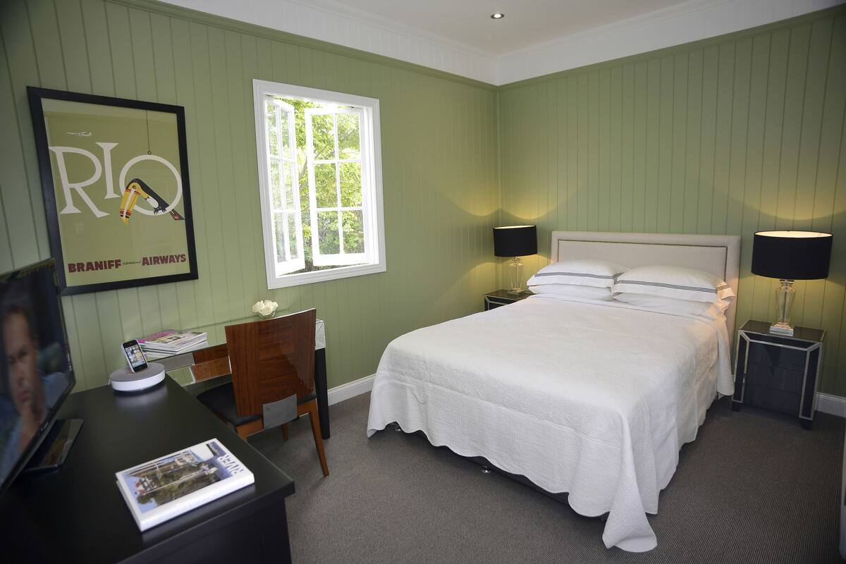 A hotel room with a bed, two bedside tables with lamps, a desk with a computer and a phone, a chair, a window, and a framed picture on the wall.