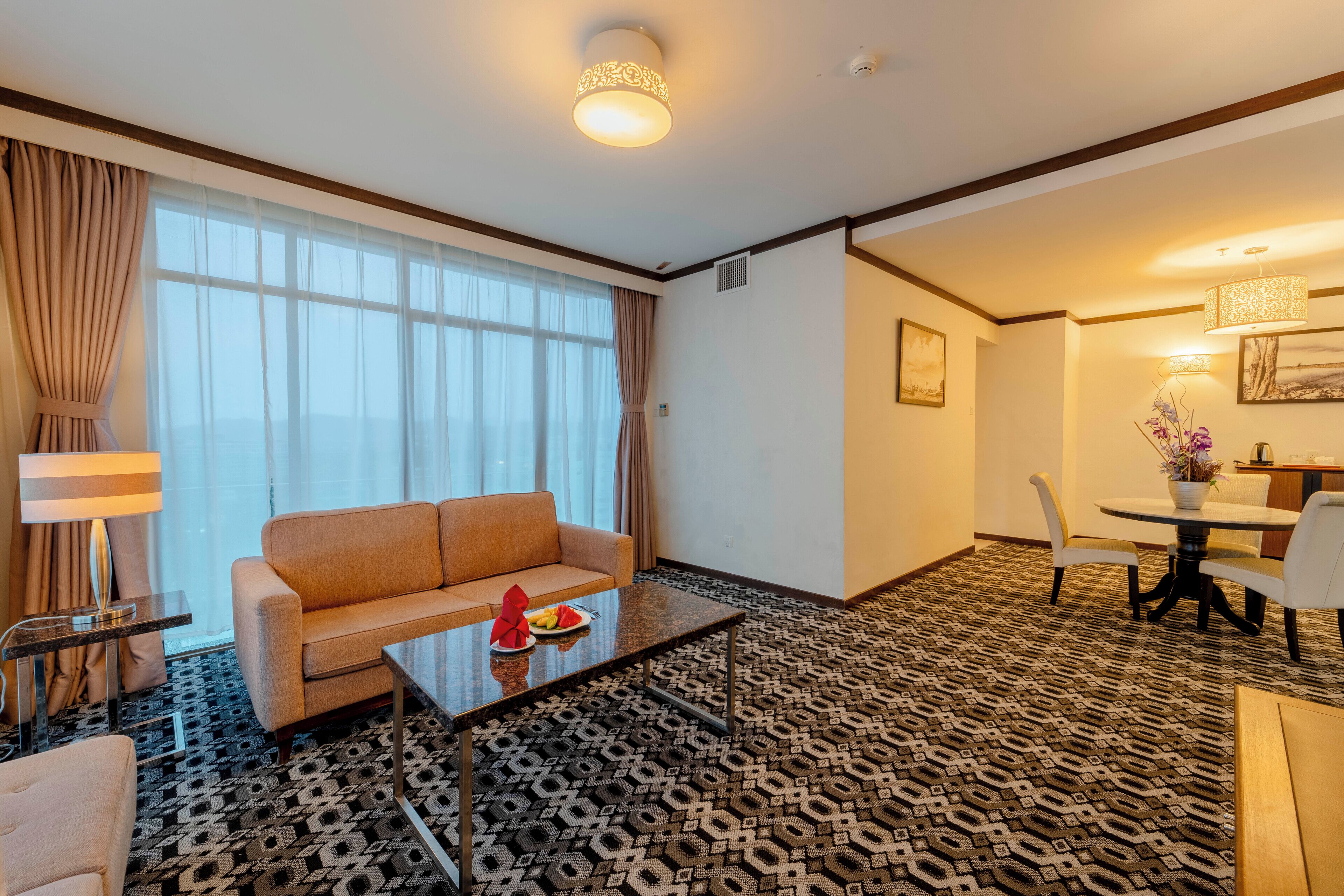 A hotel room with a sofa, coffee table, dining table with chairs, and a patterned carpet.