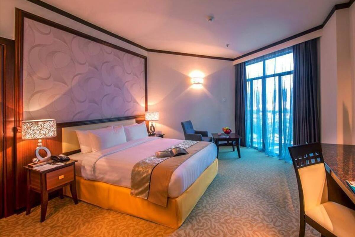 A hotel room with a large bed, a desk, a chair, a lamp, and a view of the outdoors through a window.