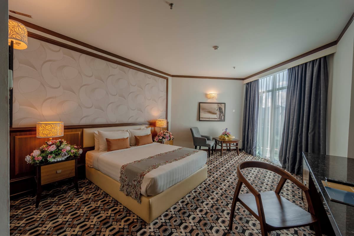 A hotel room with a large bed, a desk, a chair, a small table with a vase of flowers, and a patterned carpet.