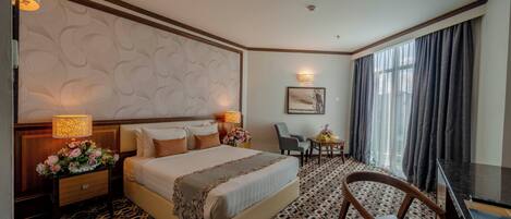 Habitación de hotel con una cama grande, un escritorio, una silla, una mesita con un jarrón de flores y una alfombra estampada.