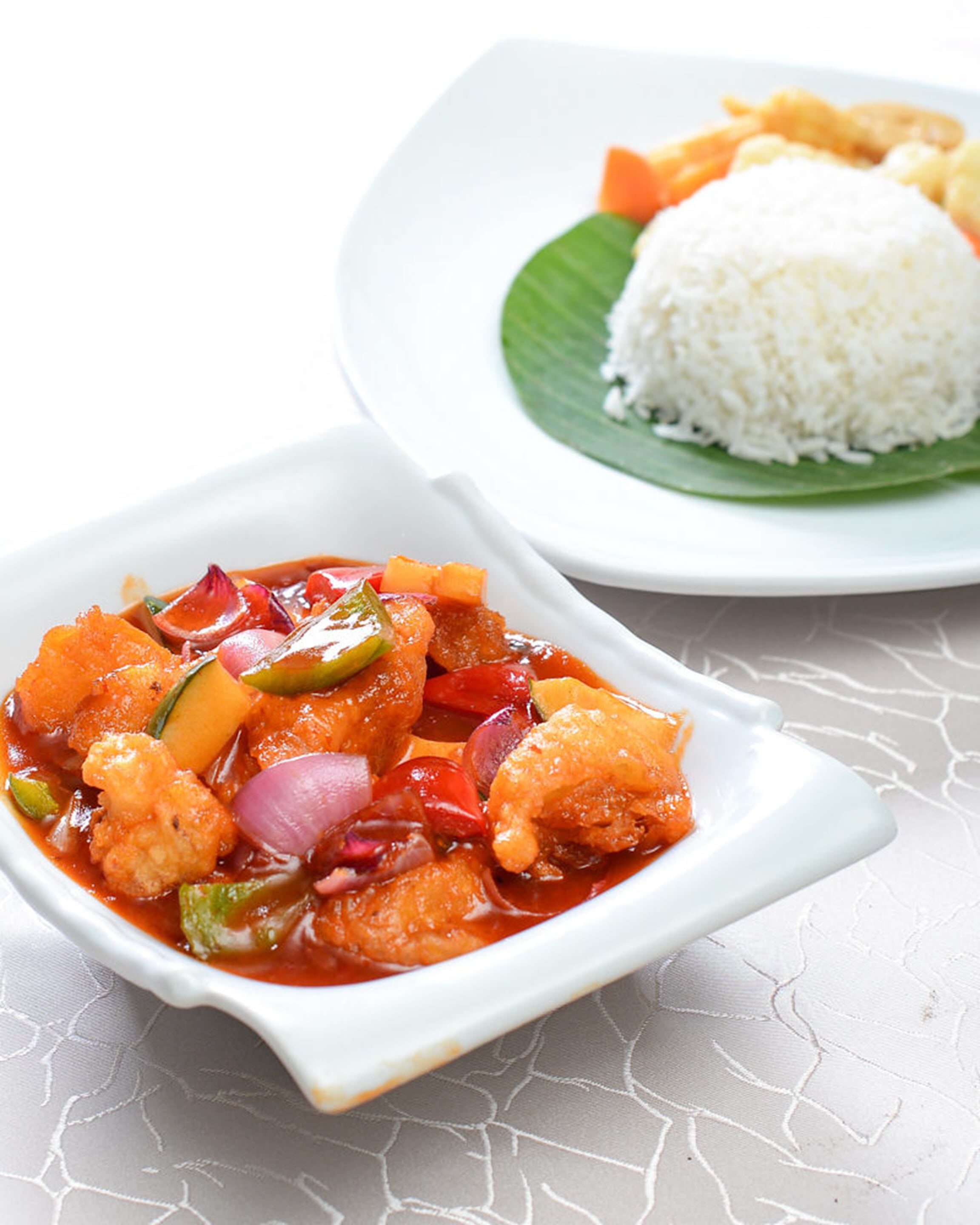 A white ceramic bowl filled with a red curry dish containing vegetables and chunks of meat, accompanied by a plate of white rice on a green leaf.