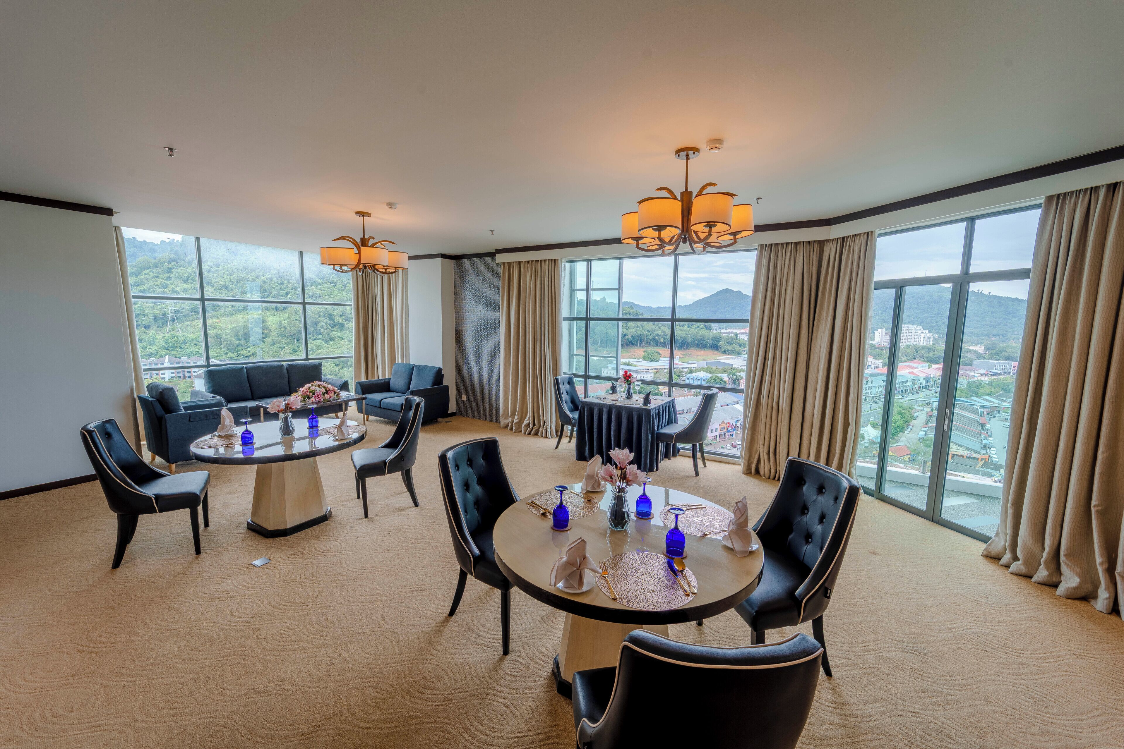 Una habitación amplia con una mesa redonda dispuesta para comer, varias sillas, un sofá y grandes ventanales que ofrecen vistas a la ciudad y a las montañas.