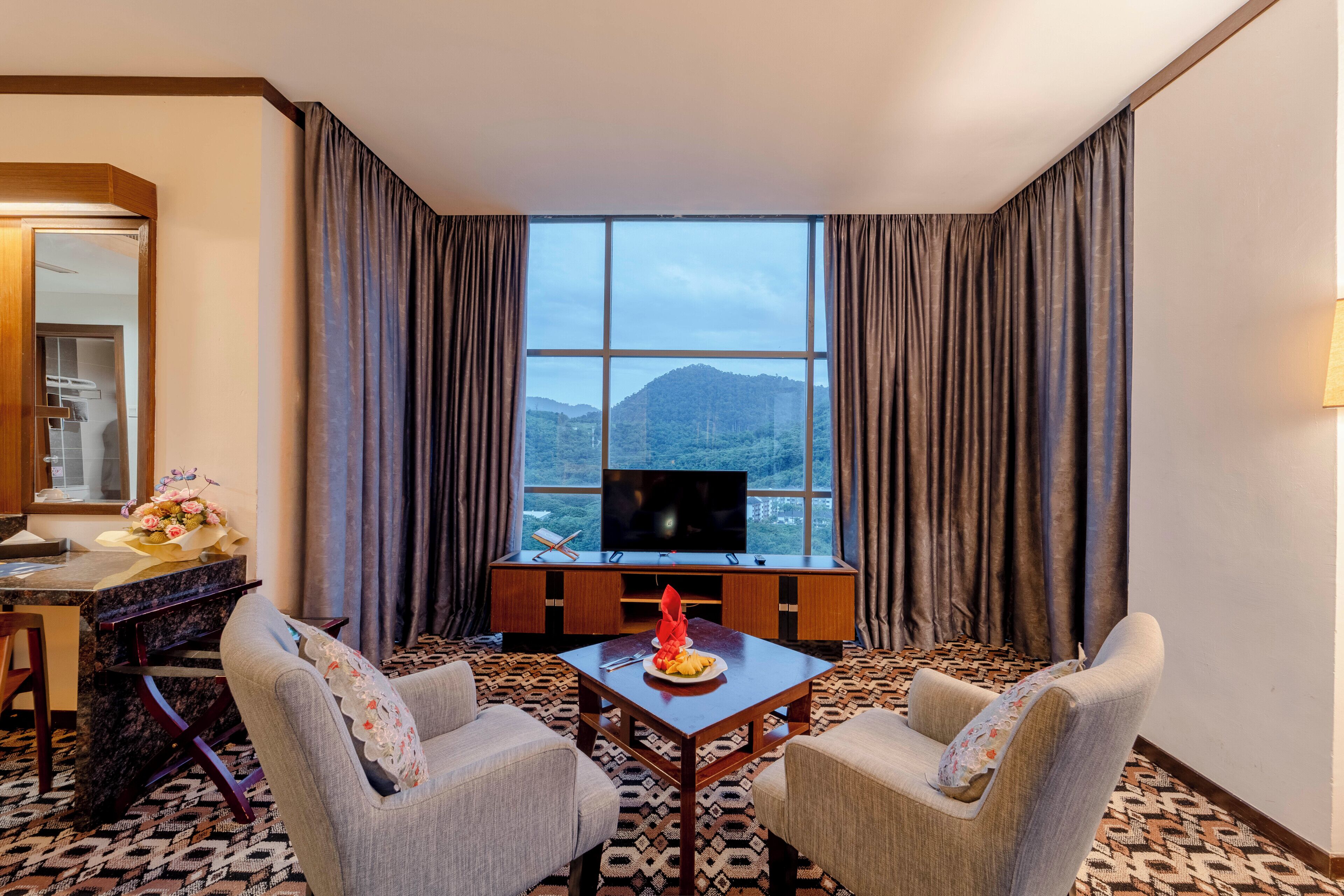 Une chambre d’hôtel avec une grande fenêtre, une télévision, une table avec une assiette de fruits, deux fauteuils et une moquette à motifs.
