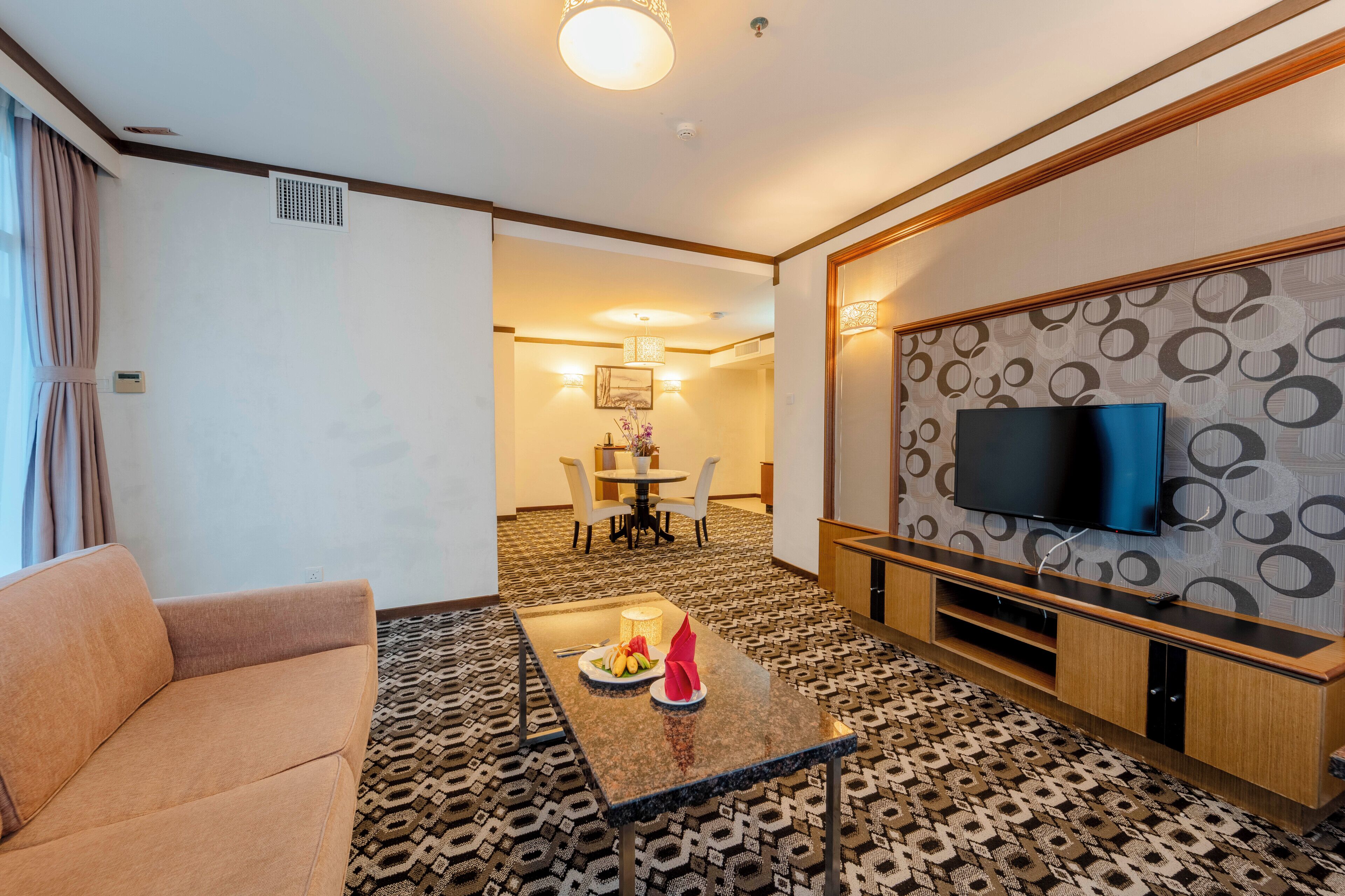 A hotel room with a sofa, a coffee table with a fruit plate, a TV on the wall, and a patterned carpet.