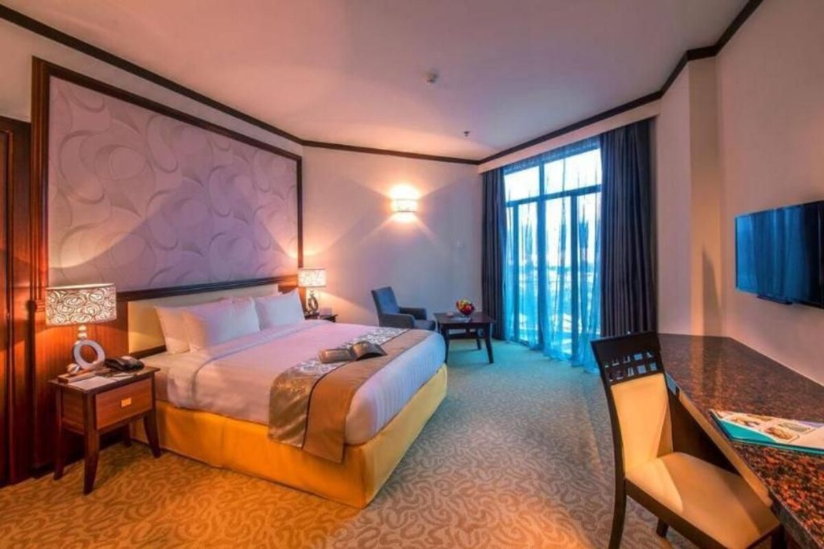 A hotel room with a large bed, a desk with a chair, a TV on the wall, and a window with curtains.