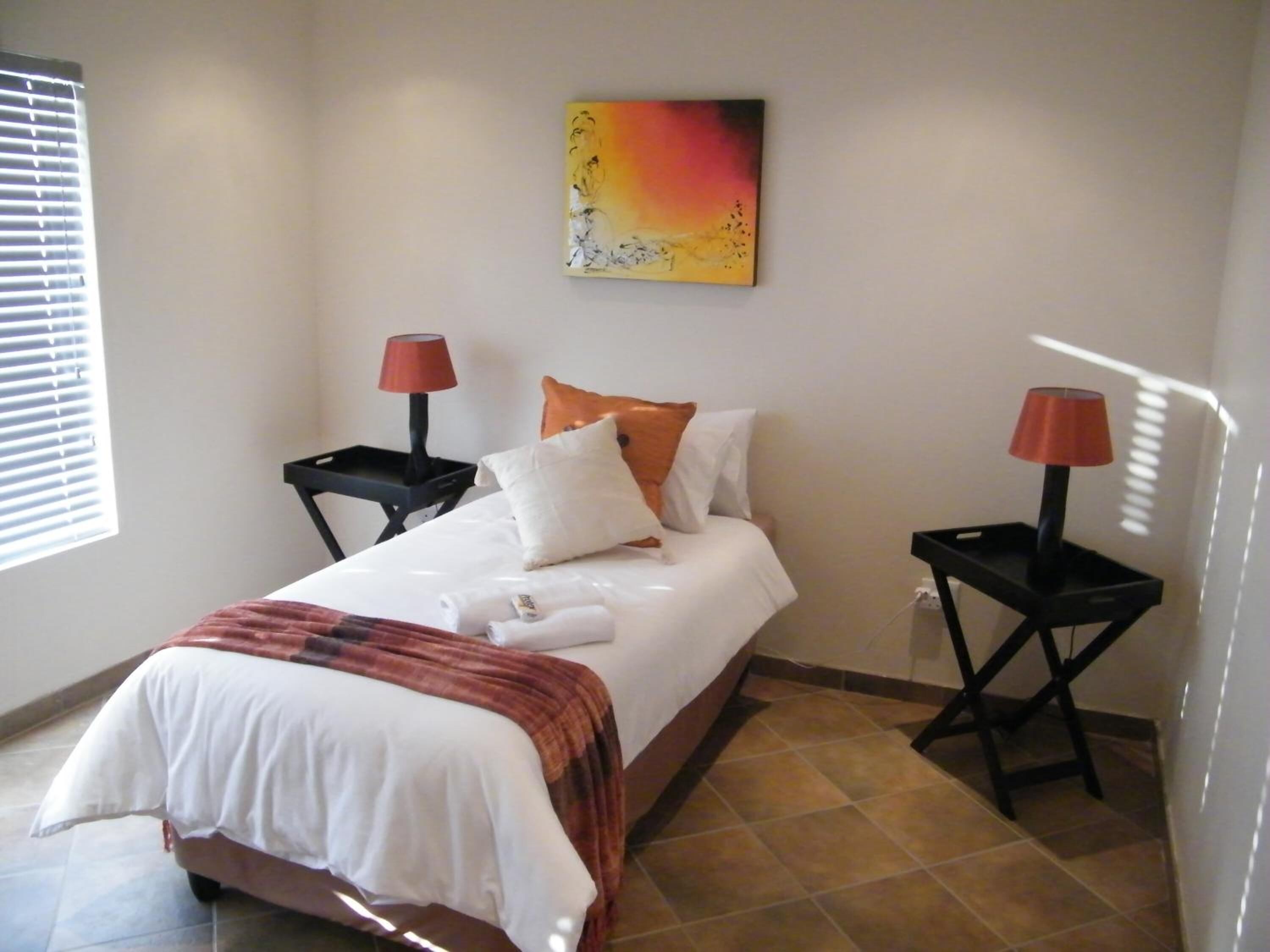 Una cama bien hecha con ropa de cama blanca y una manta roja, dos mesitas de noche con lámparas, un cuadro en la pared y una ventana con persianas.