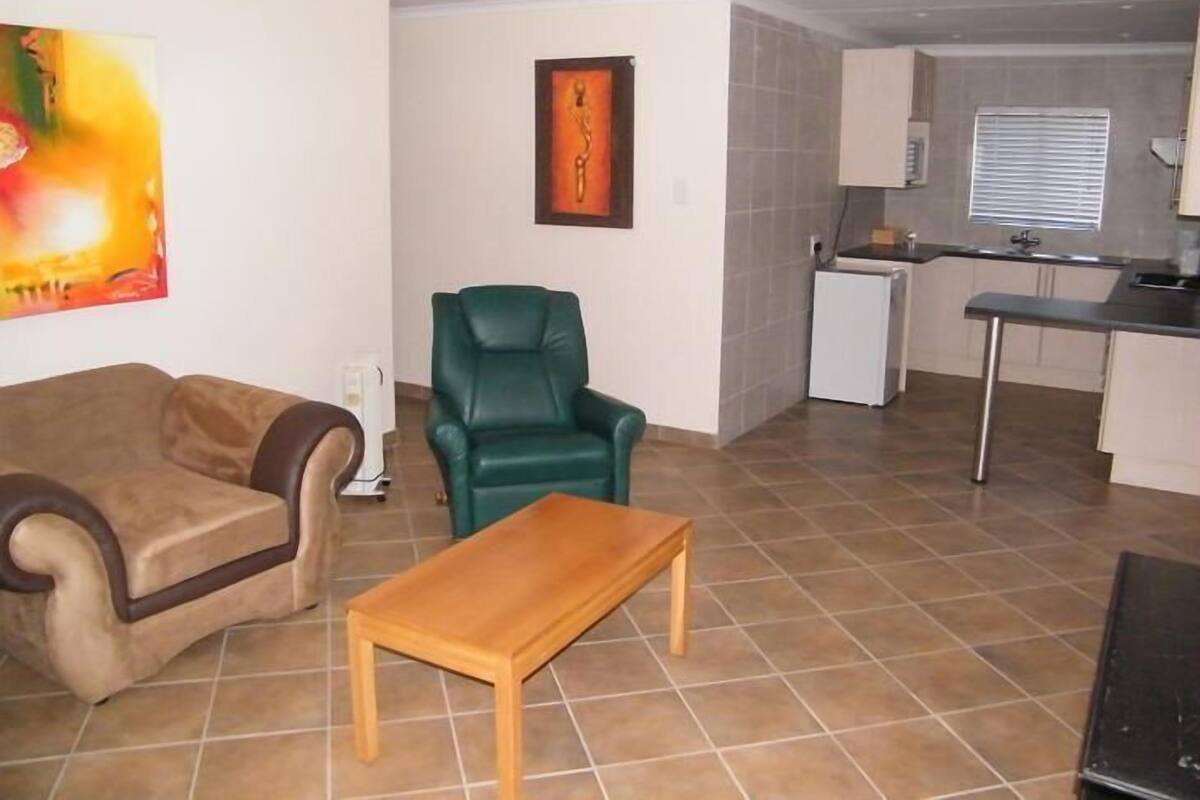 A living room with a sofa, a green chair, a wooden table, a painting, and a kitchen area with a refrigerator and sink.