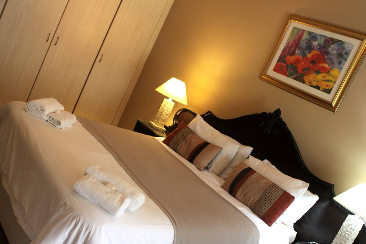 A hotel room with a neatly made bed, a bedside lamp, a framed floral painting on the wall, and a wardrobe.