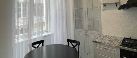 Uma cozinha com uma mesa redonda, cadeiras, fogão e uma janela com cortinas.