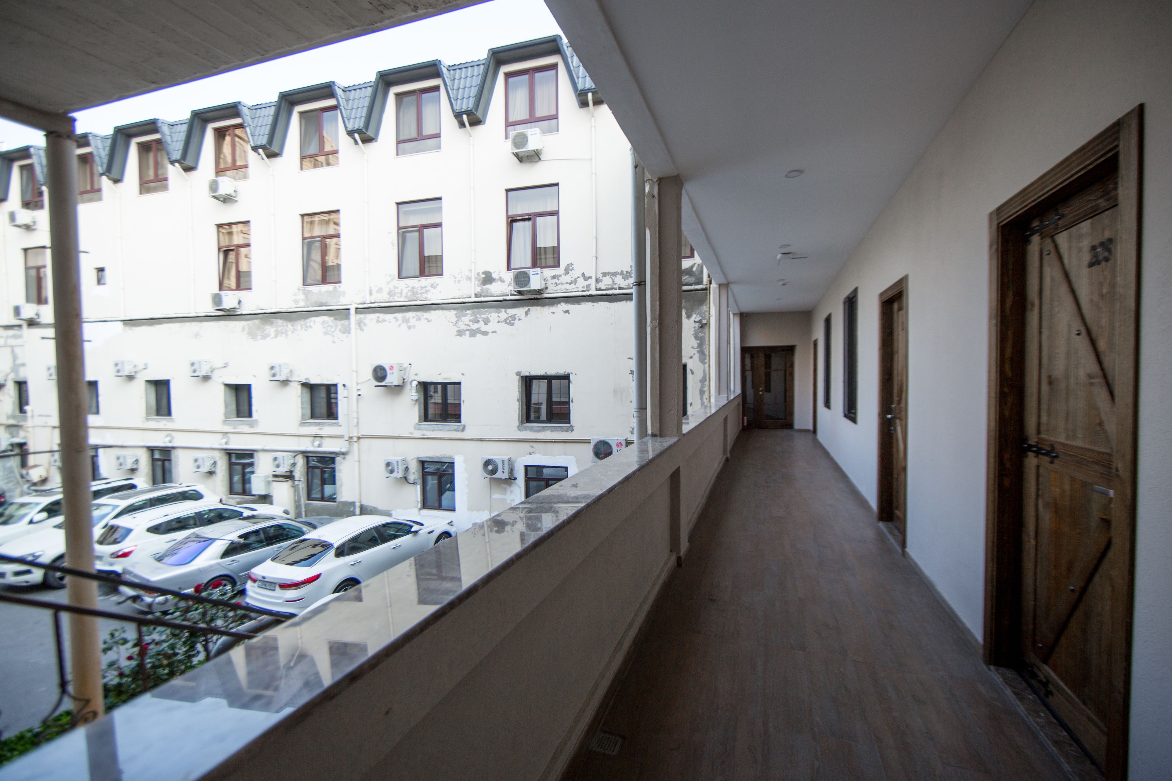 A corridor with a wooden floor, a balcony with a glass railing, and a view of parked cars and buildings in the background.