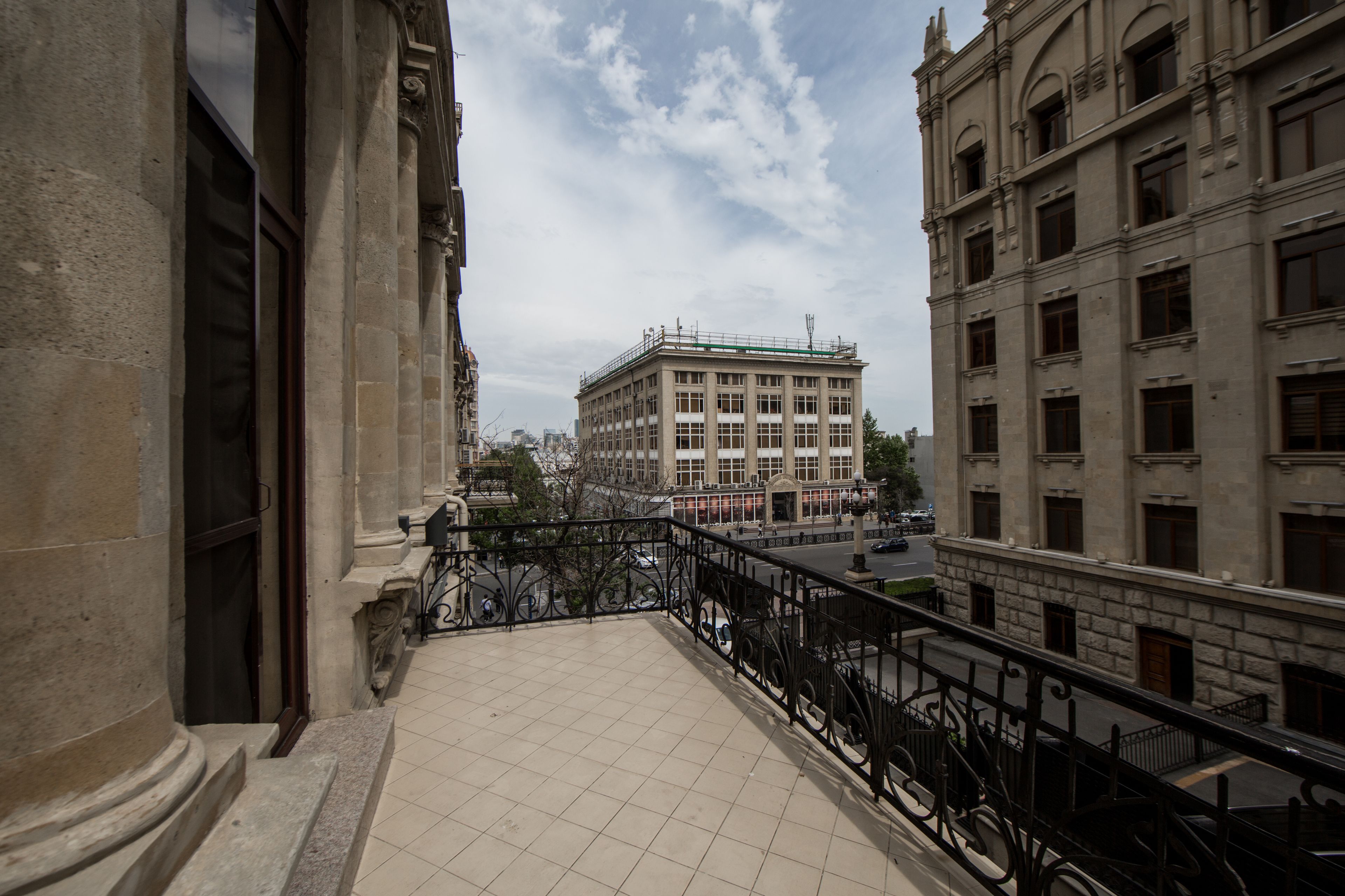 A balcony with a tiled floor and black wrought iron railing, overlooking a street with buildings and a clear sky.