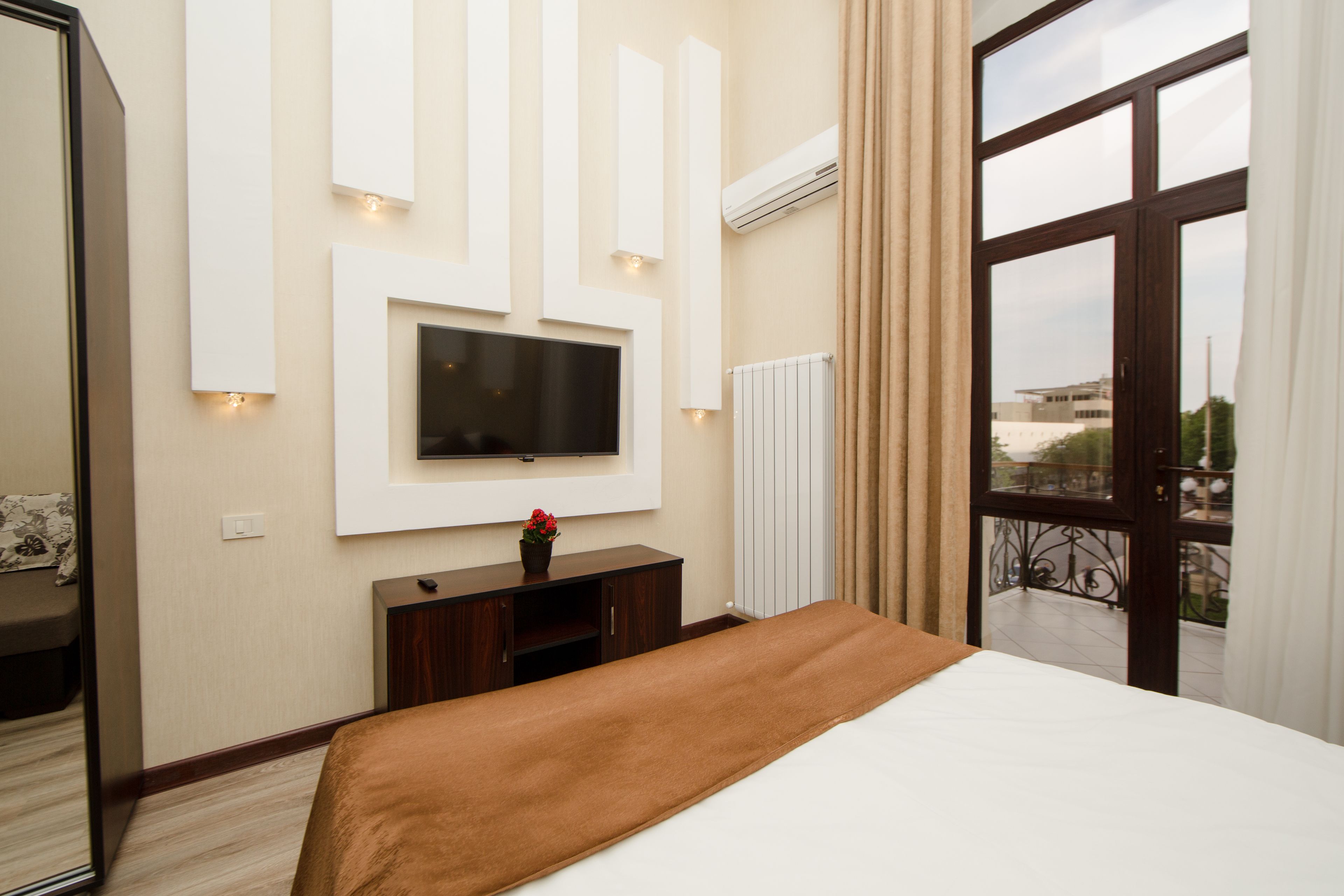A hotel room with a large bed, a TV mounted on the wall, a small wooden cabinet, and a window with a view of the city.