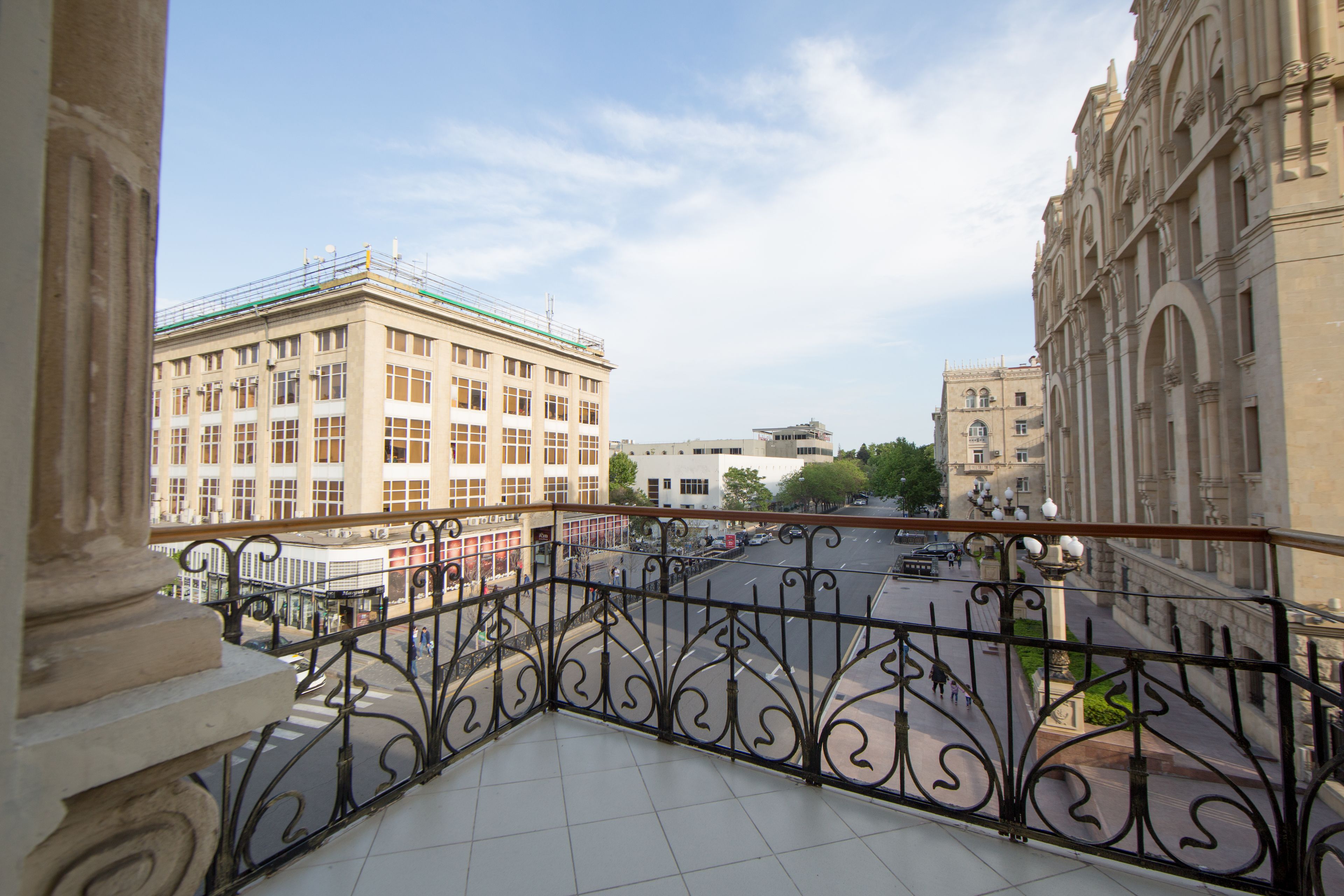 A balcony with ornate black iron railings overlooking a street with buildings and a clear sky.