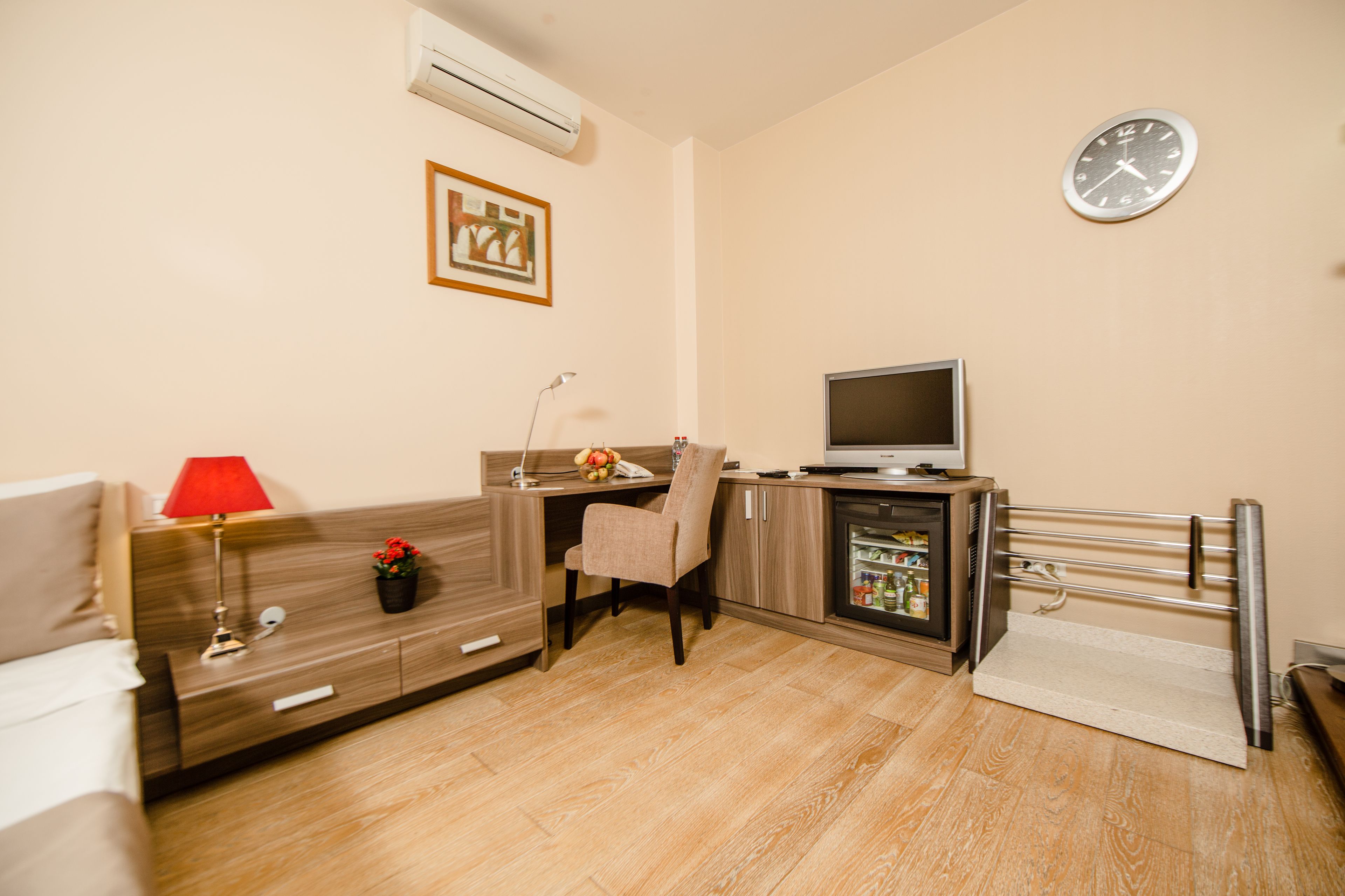 A hotel room with a bed, a desk with a computer, a chair, a TV, a clock, and a small refrigerator.