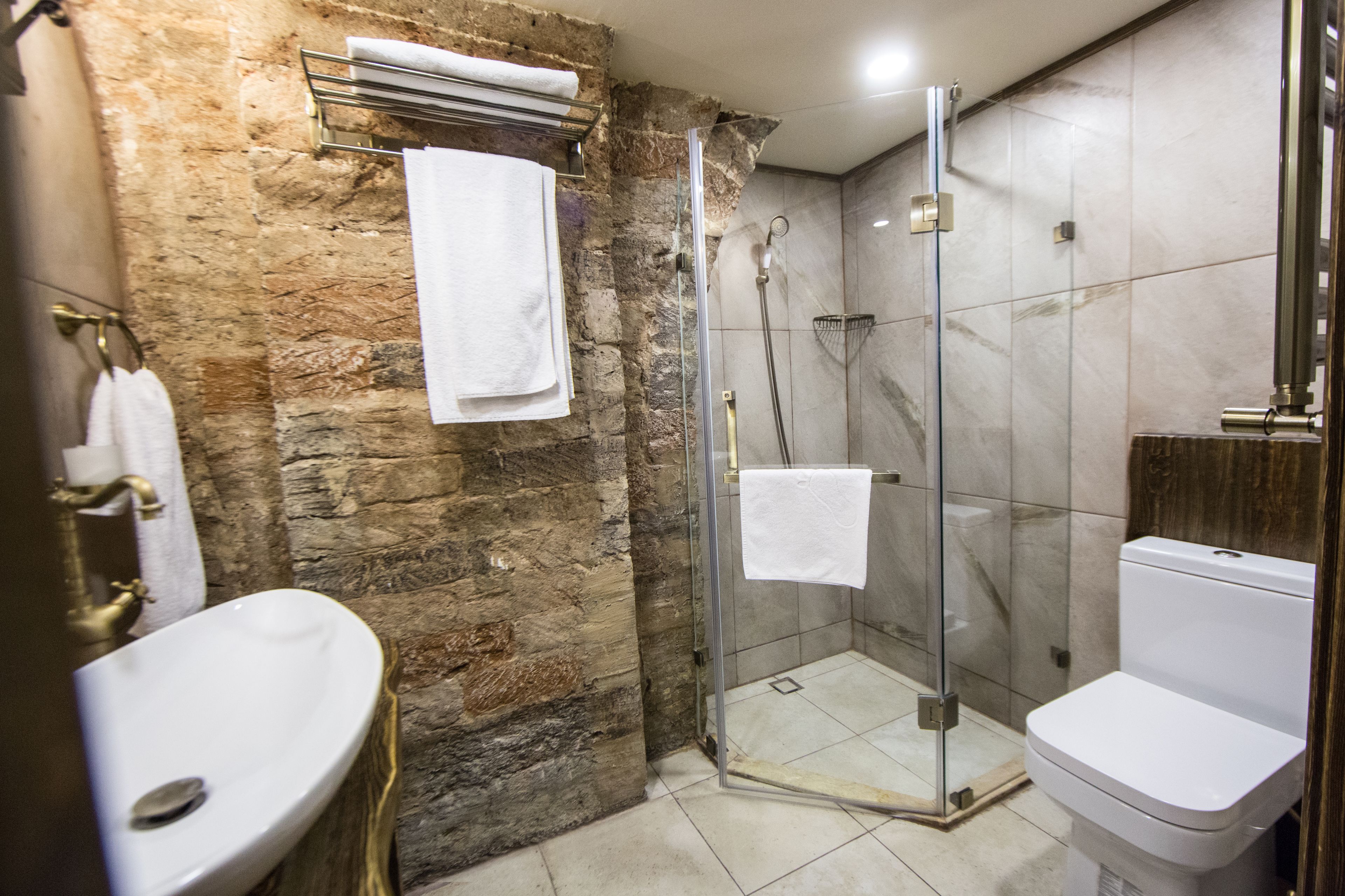 A bathroom with a stone wall, a glass-enclosed shower, a white sink, and a toilet.