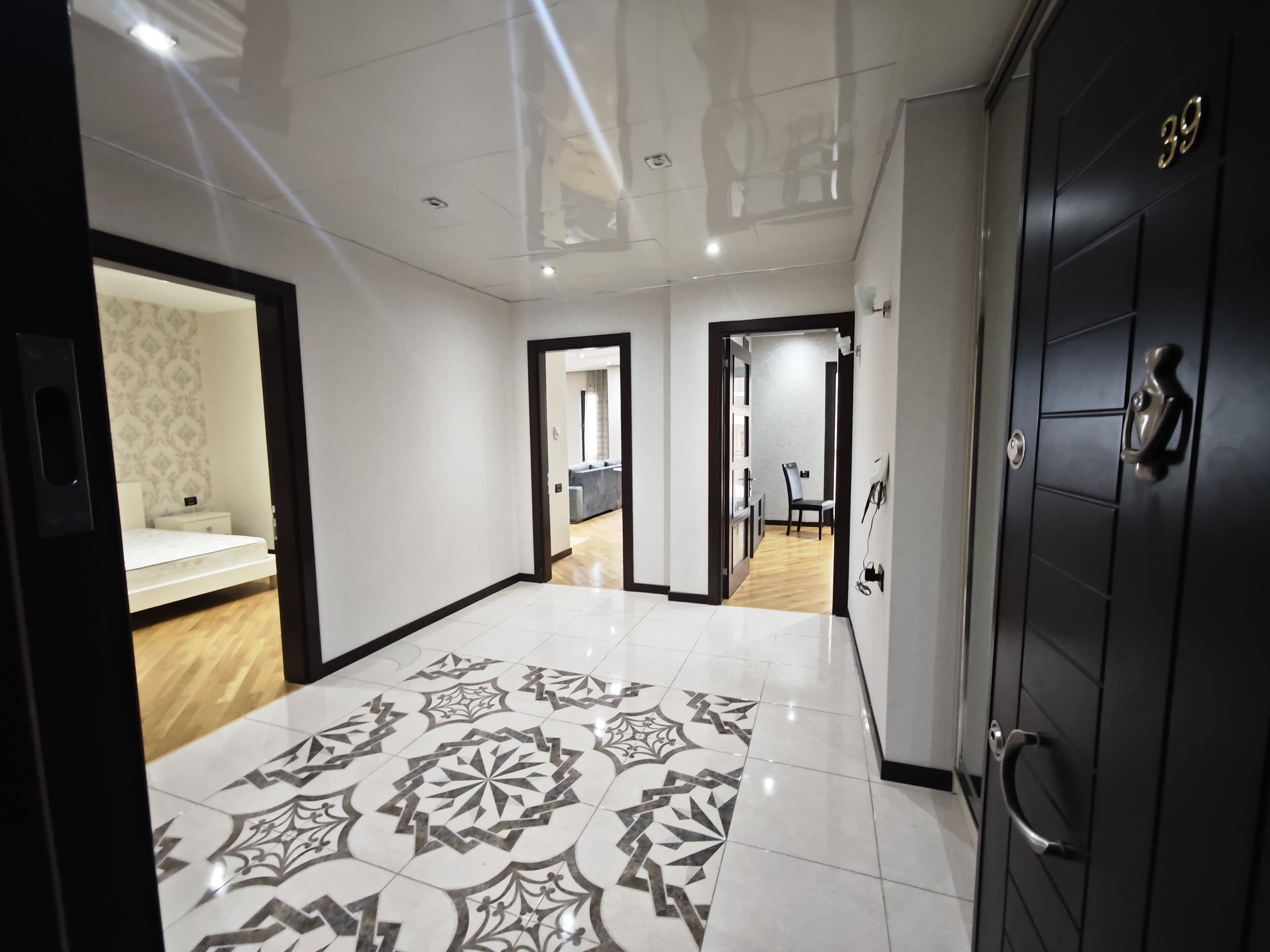 A spacious hallway with a patterned floor, a dark door with a number, and a glimpse of a room with a bed and a patterned wall.
