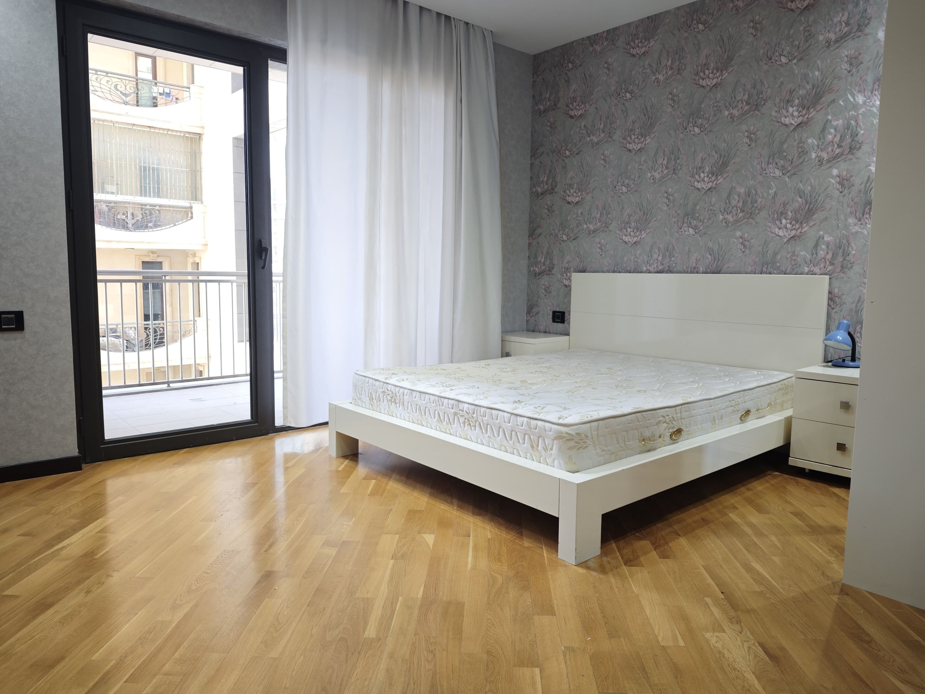 Ein Schlafzimmer mit einem weißen Bett, Holzboden, einem Nachttisch, einem Schreibtisch und Blick auf einen Balkon.