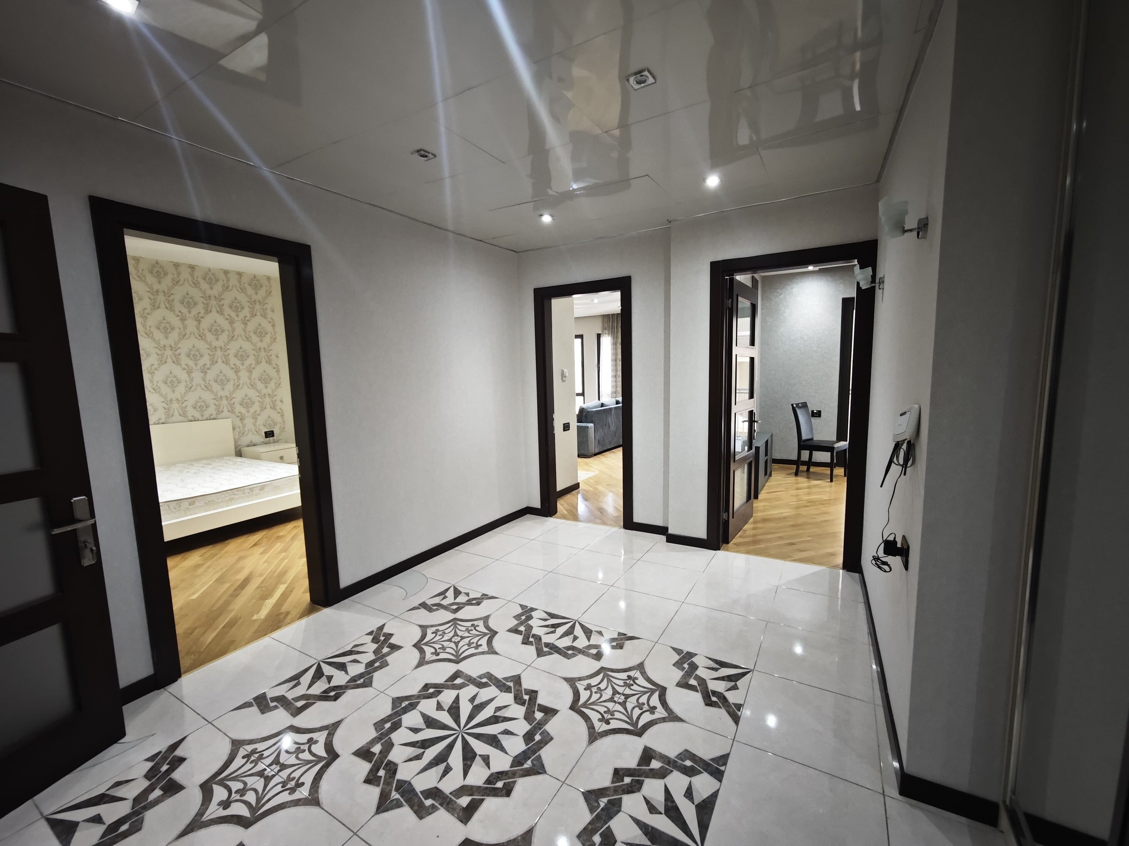 A spacious hallway with patterned floor tiles, a bed with a patterned headboard, and a seating area with a chair and a table.