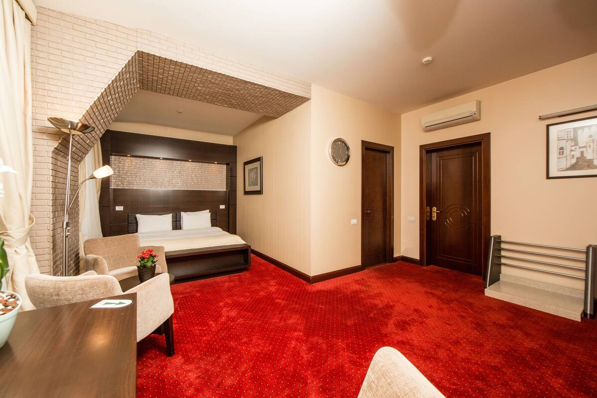 Ein Hotelzimmer mit einem großen Bett, einem Schreibtisch mit Stuhl, einem kleinen Tisch mit Pflanze, einem roten Teppich, einer Ziegelwand als Gestaltungselement, einer Uhr und einer Tür.