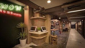 Interno di un ostello con un cartello al neon, una reception con computer e un corridoio che porta alle camere.