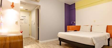 A hotel room with a bed, a desk, an air conditioning unit, and a door leading to another room.
