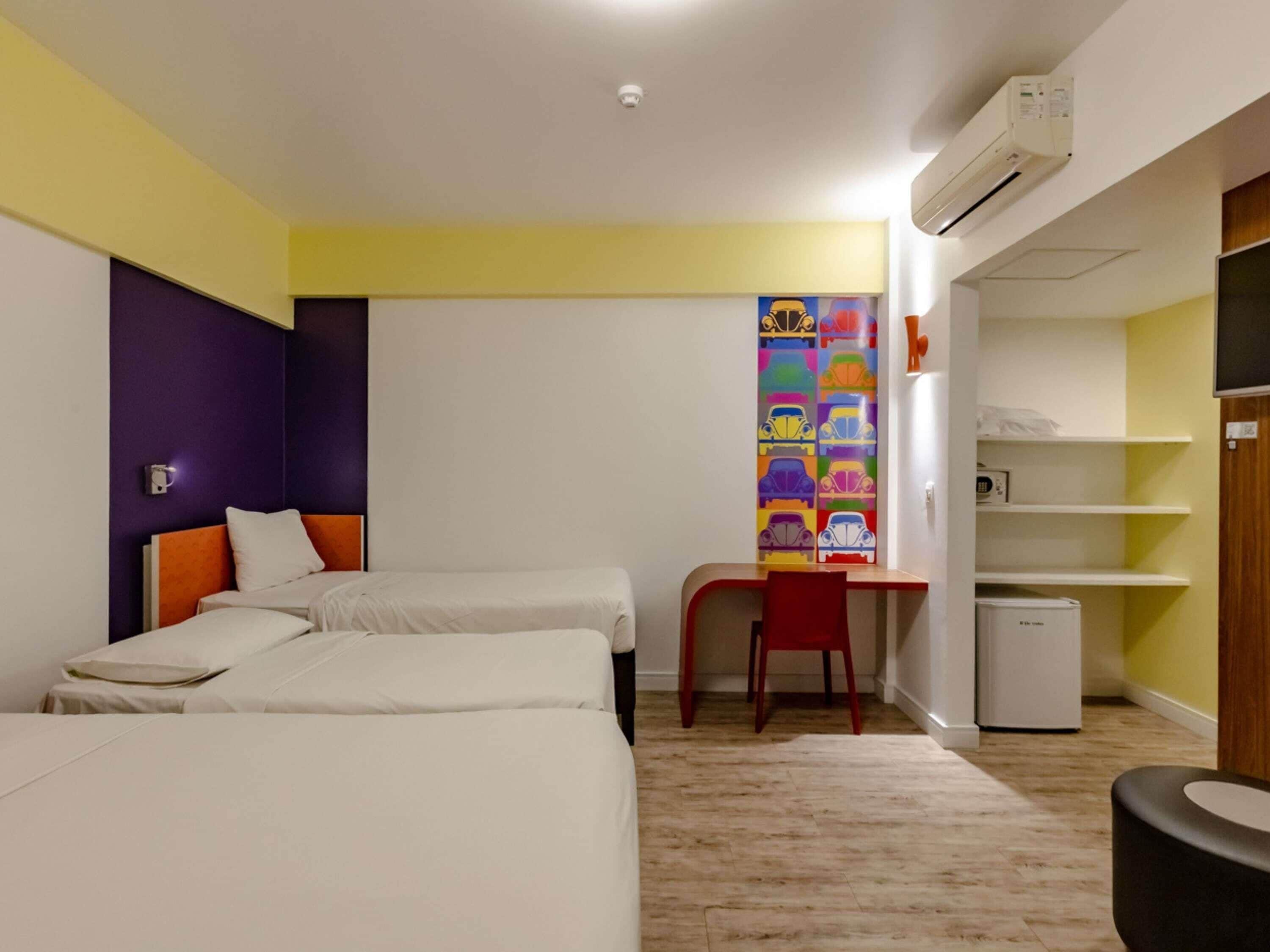A hotel room with two beds, a desk, a chair, a refrigerator, and a wall-mounted air conditioner.