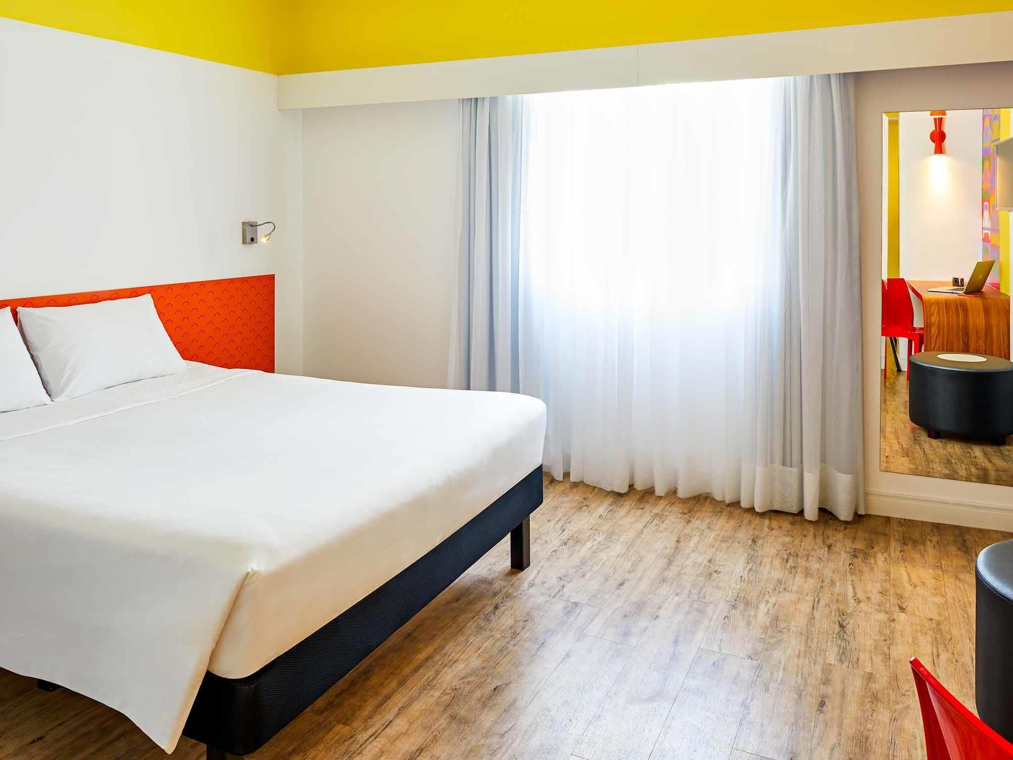 A hotel room with a bed, a window with curtains, a wooden floor, and a small table with a chair.