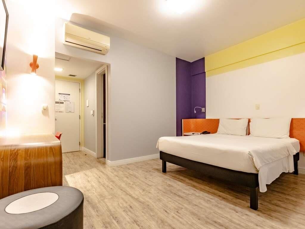 A hotel room with a bed, a desk, and an air conditioning unit.