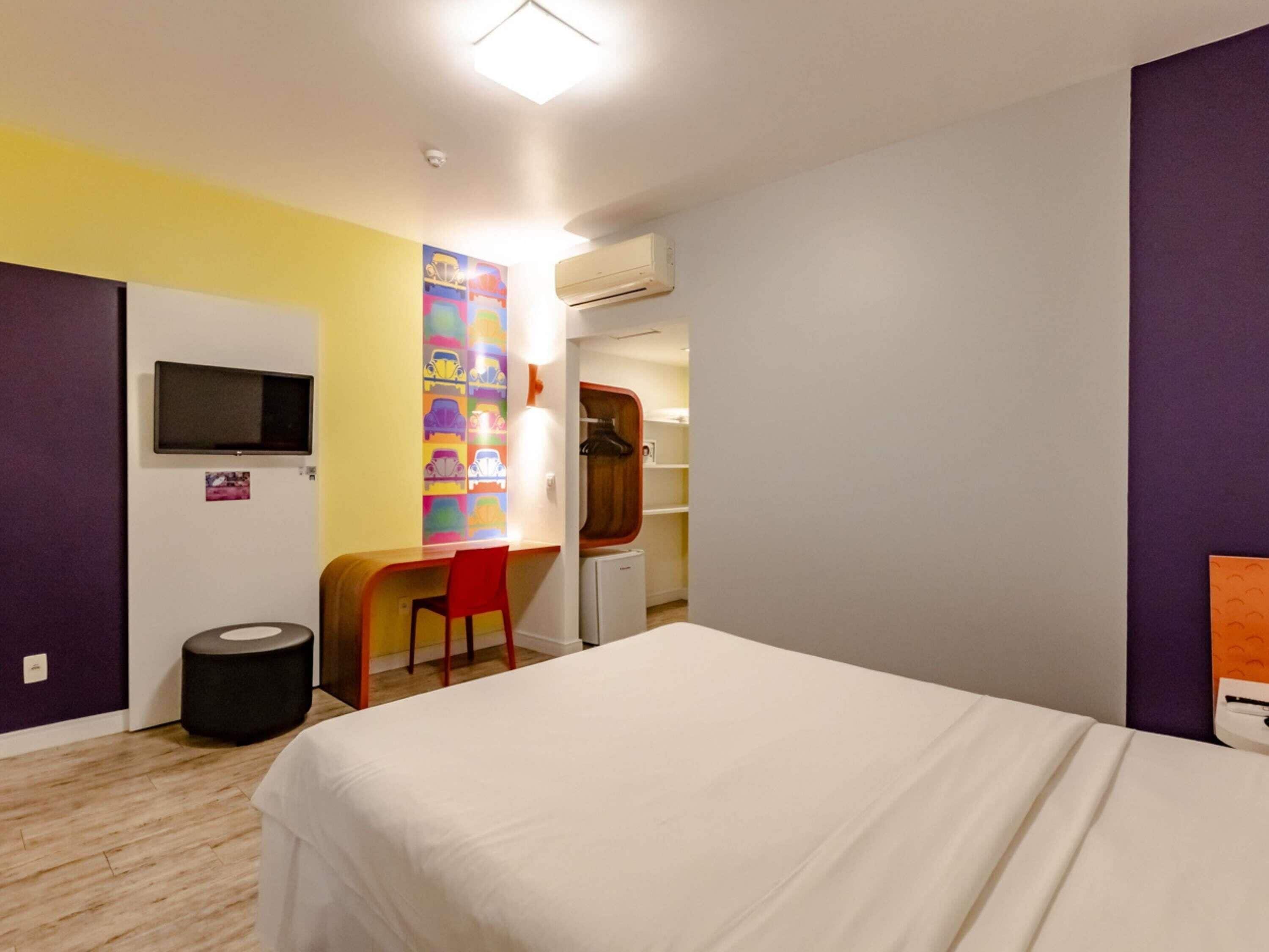 A hotel room with a bed, a desk, a chair, a television, and a colorful wall decoration.