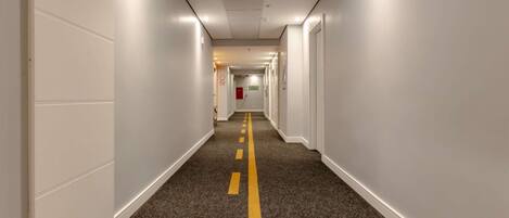 Un pasillo de hotel con pisos alfombrados, paredes blancas y una línea amarilla doble delimitando.