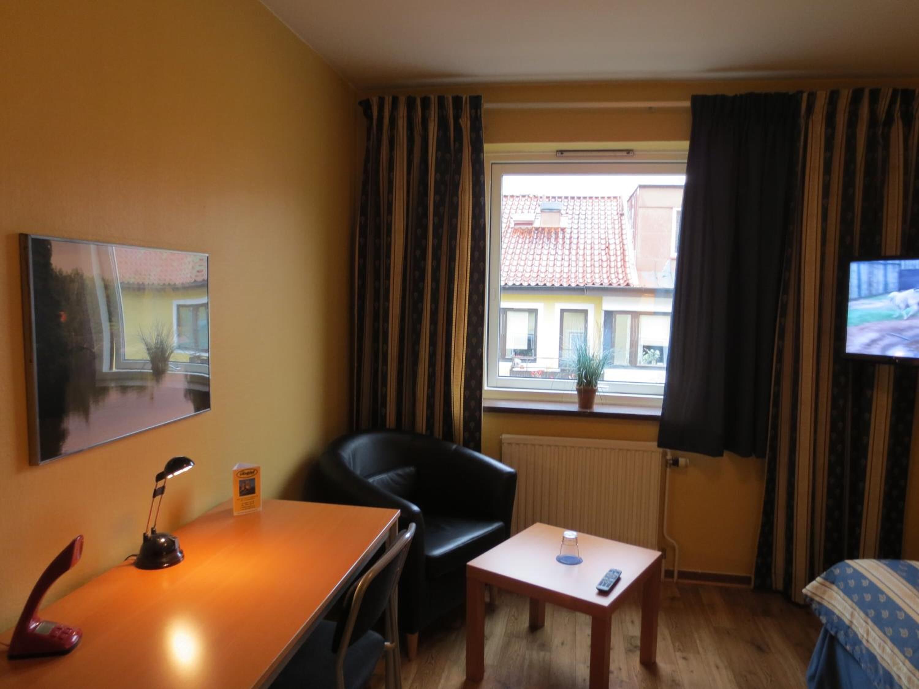 A hotel room with a bed, a desk with a chair, a small table, a TV, and a window with curtains.