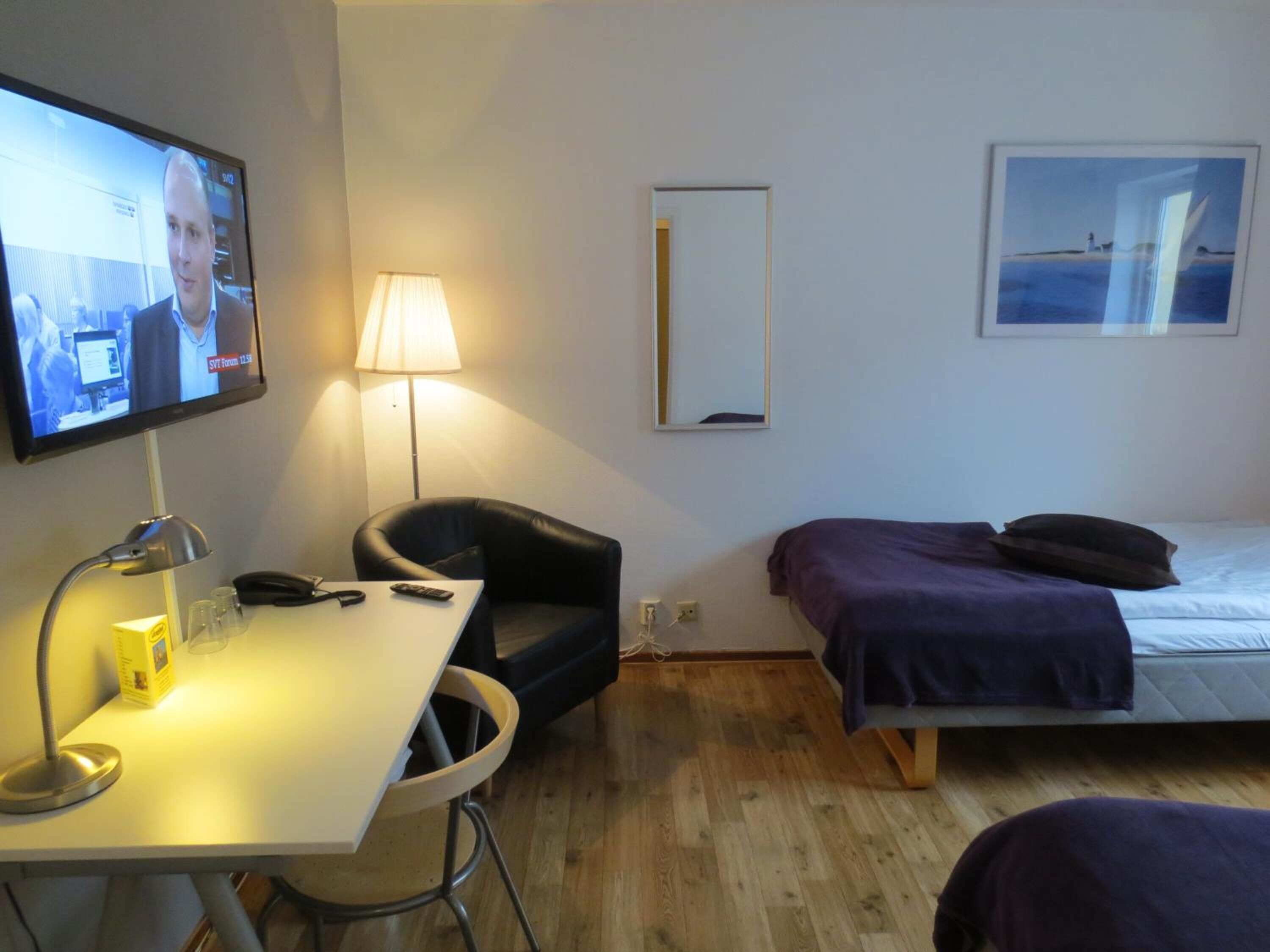 Ett hotellrum med en TV, ett skrivbord med en lampa, en stol, en säng med ett lila överkast och en inramad tavla på väggen.