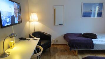 Ett hotellrum med en TV, ett skrivbord med en lampa, en stol, en säng med ett lila överkast och en inramad tavla på väggen.