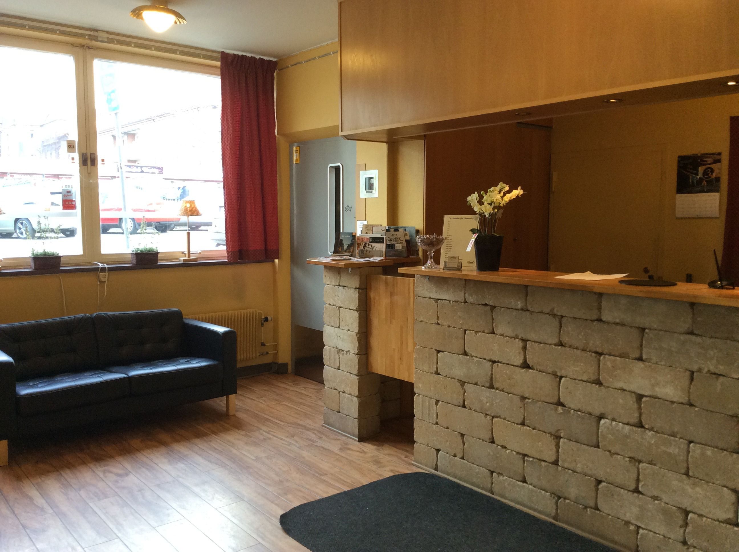 A cozy hotel lobby with a black sofa, a brick reception desk, and a window with a view of the street.