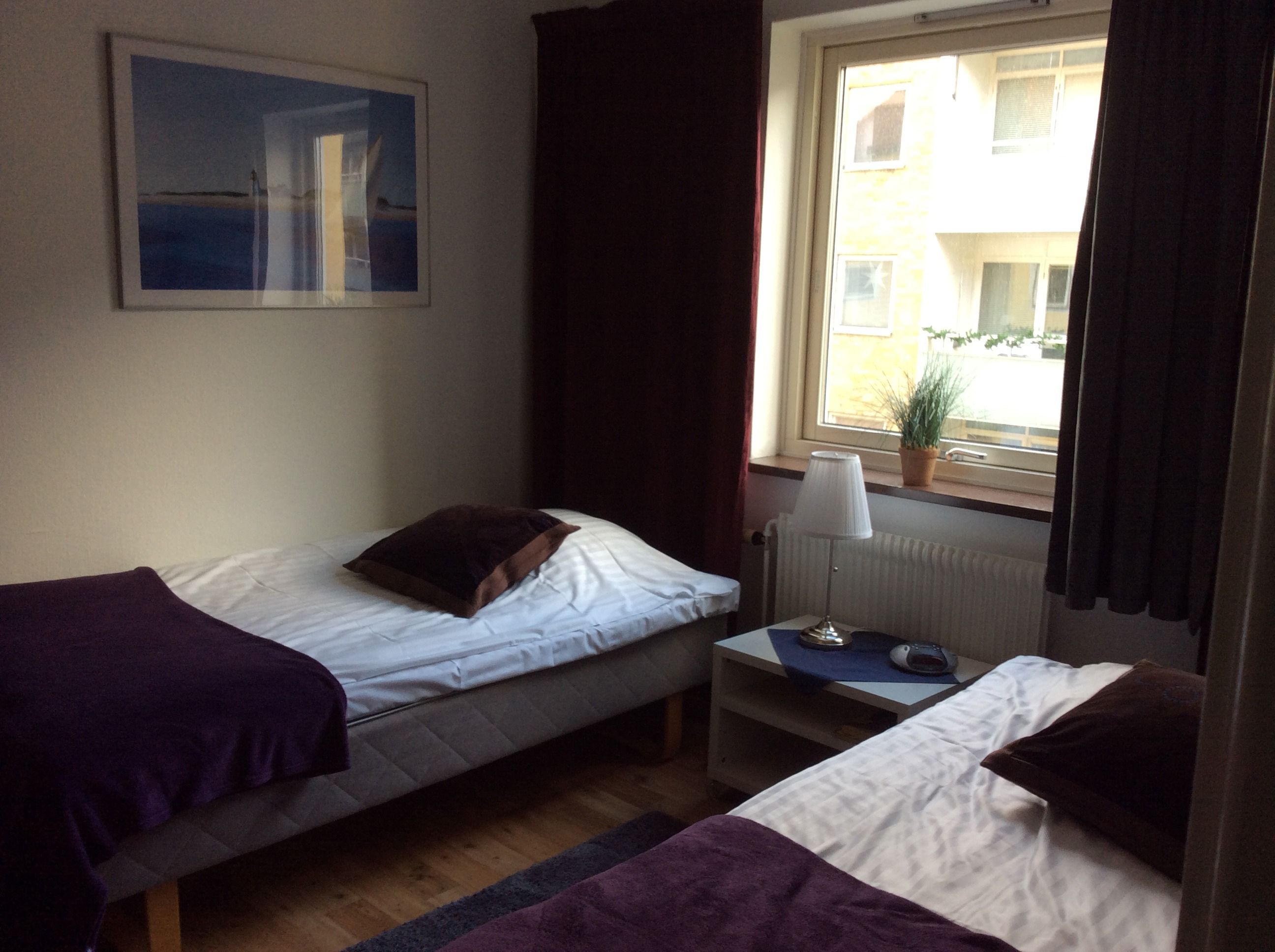 A room with two beds, a small table with a lamp, a window with curtains, and a painting on the wall.