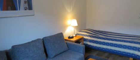 A room with a grey sofa, a bed with a blue and white striped bedspread, a wooden nightstand with a lamp, and a framed picture of lighthouses on the wall.