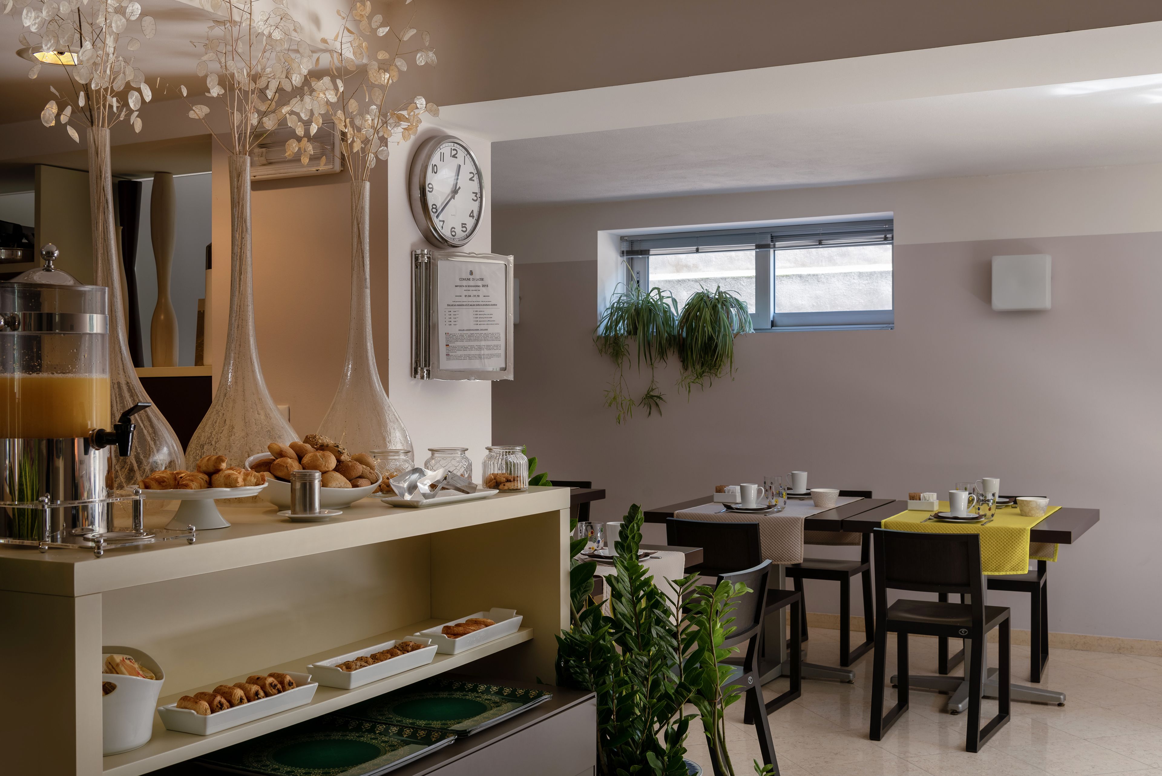 A breakfast area with a buffet setup, including pastries and a coffee machine. There is a clock on the wall and a window with plants.
